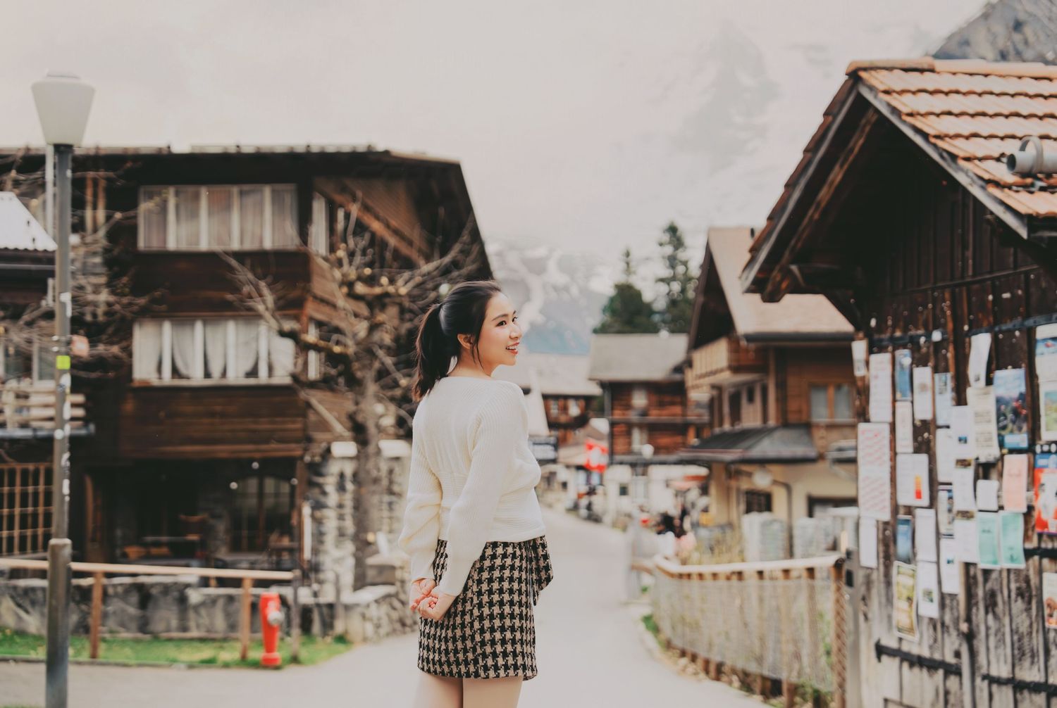 Young woman in white sweater and plaid skirt strolling along a scenic Swiss alpine village street.