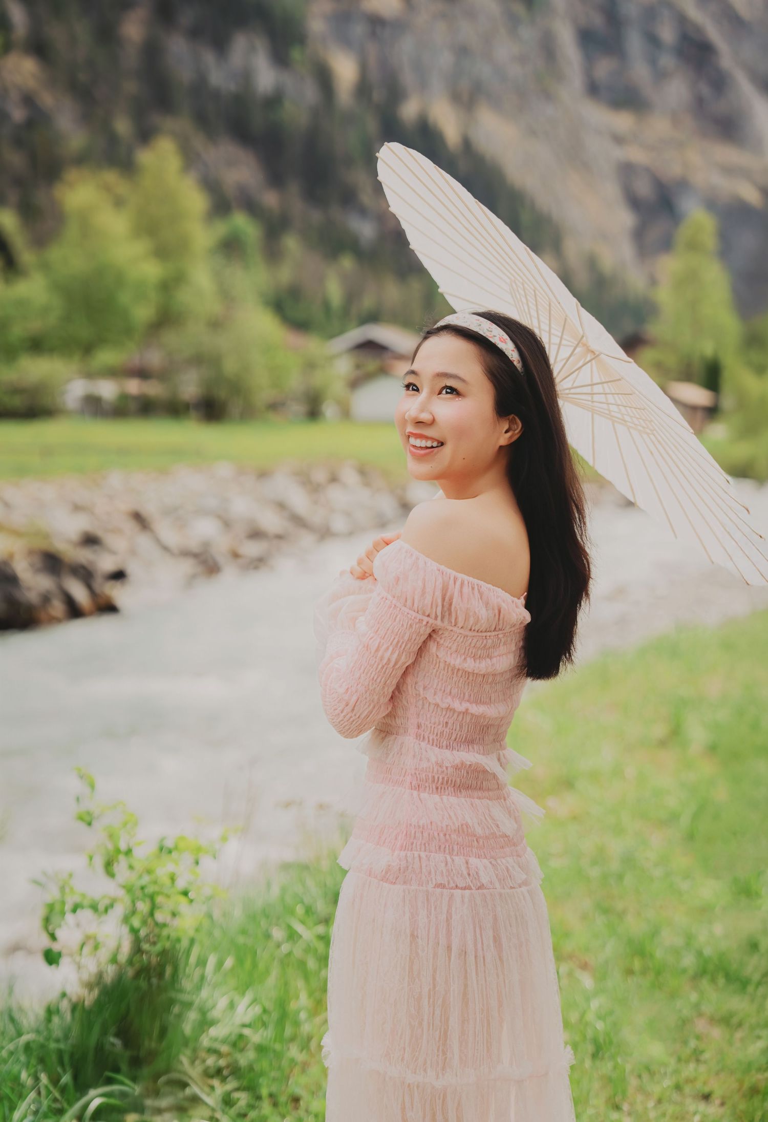 Smiling woman in off-shoulder pink dress holding white parasol beside a scenic mountain river.