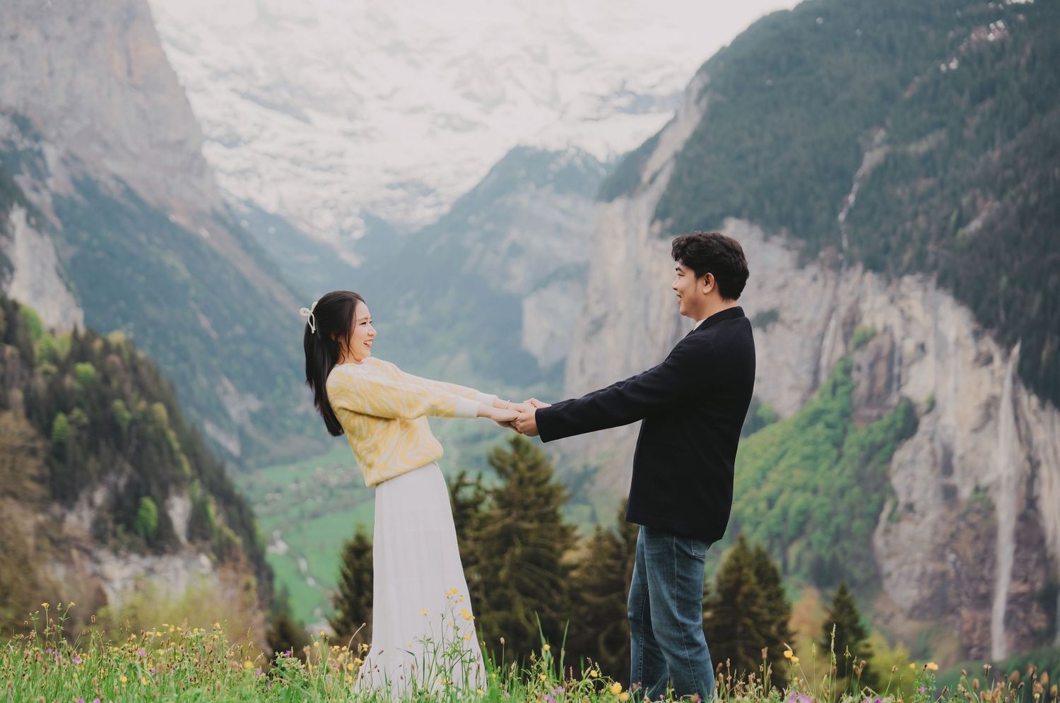 Couple holding hands facing each other in Swiss alpine meadow with dramatic mountain valley backdrop.