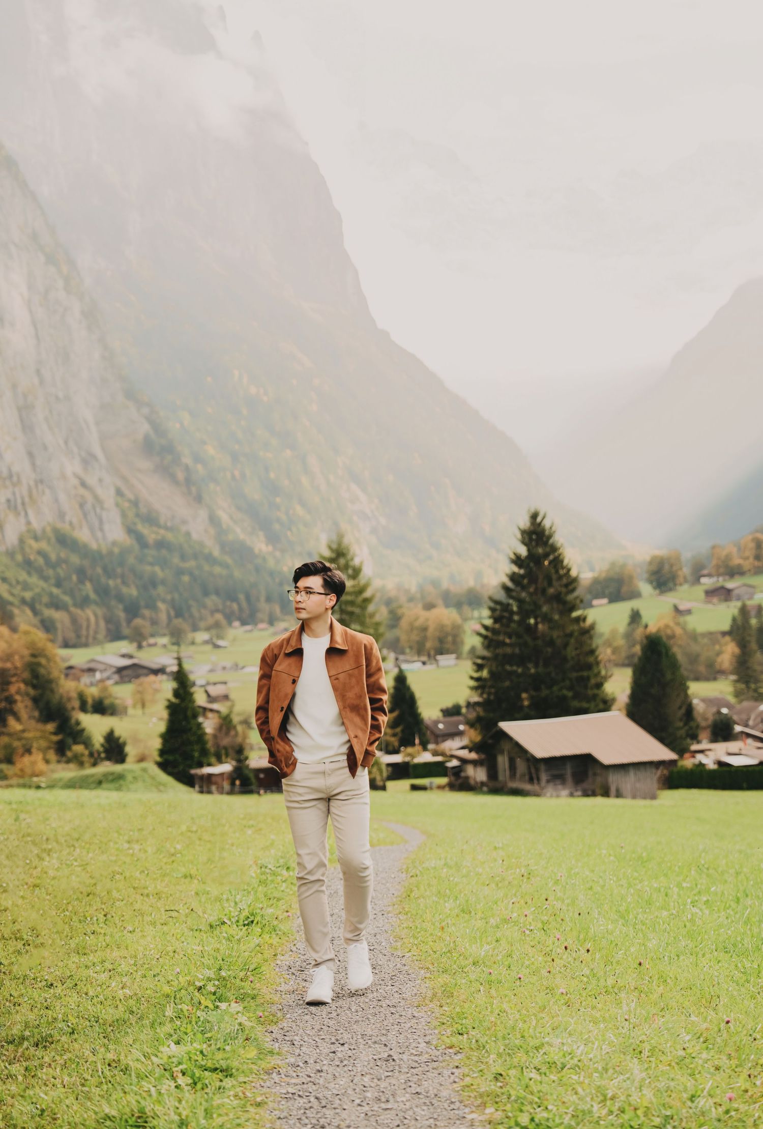 Stylish young man walking on a path through a lush Swiss alpine valley with misty mountain peaks behind.