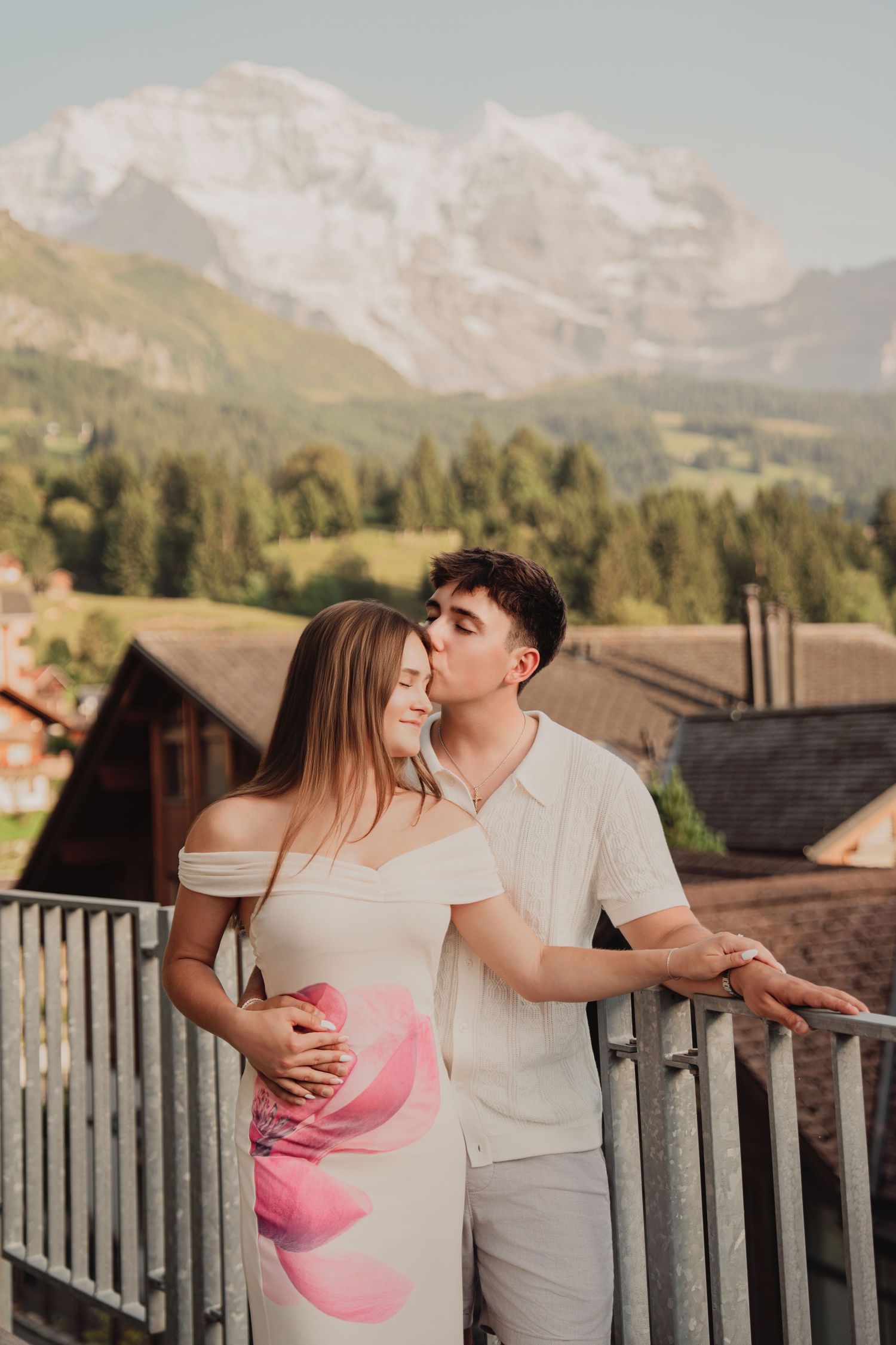 Man kissing woman on cheek while embracing on a balcony with majestic snow-covered Swiss mountain peaks behind them.