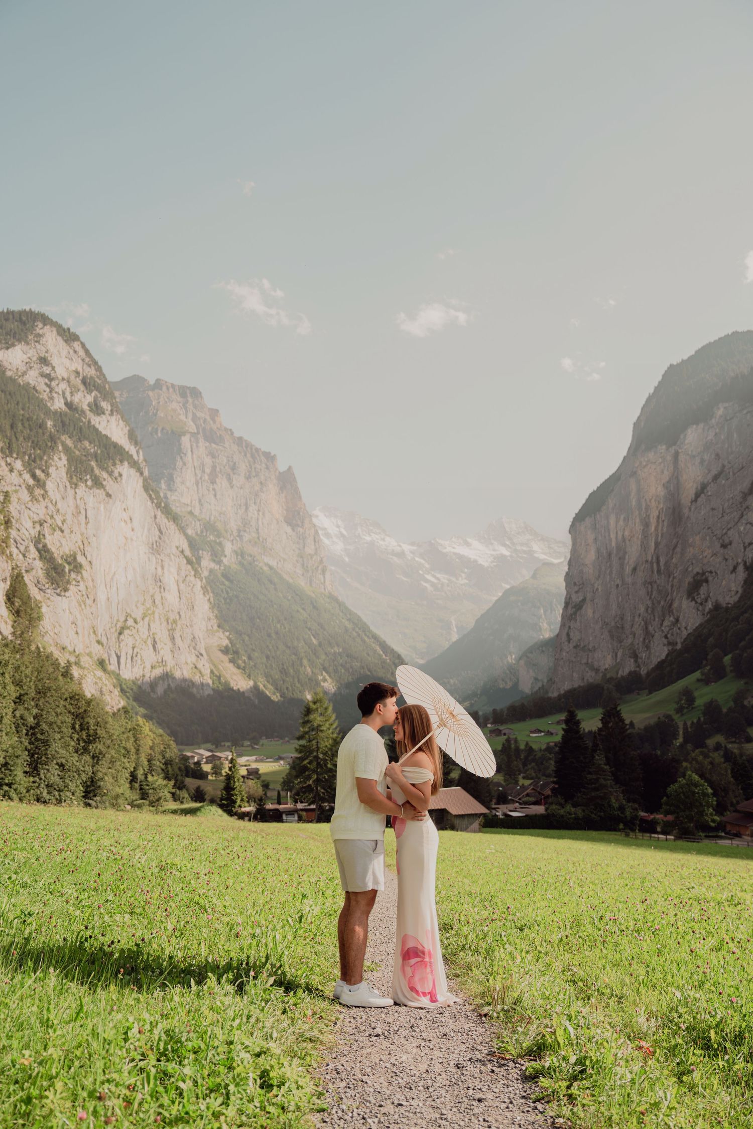 Couple kissing under white parasol on a gravel path surrounded by dramatic Swiss Alps and lush green valley.