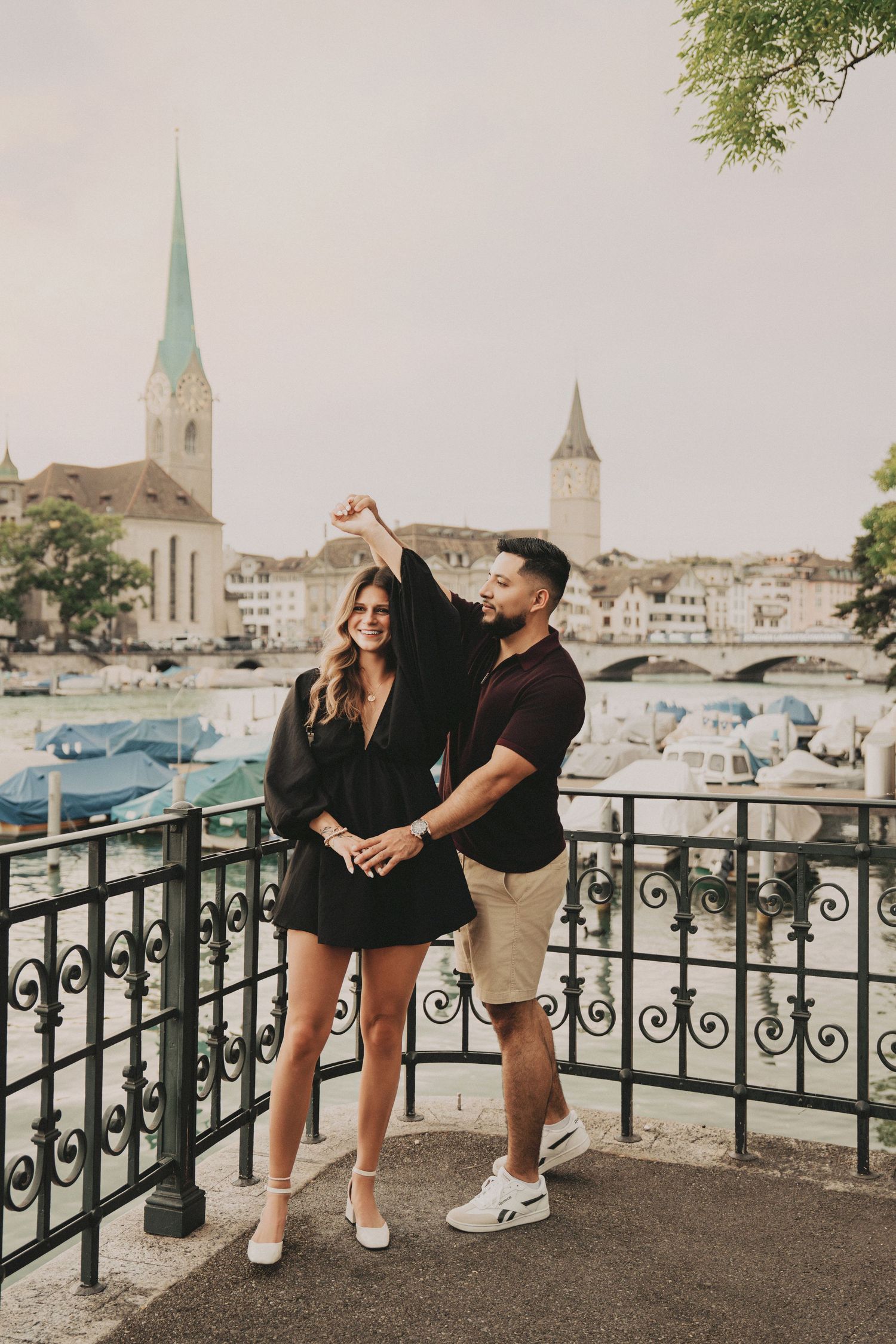 Man twirling woman in black dress on a decorative balcony with Zurich waterfront backdrop.