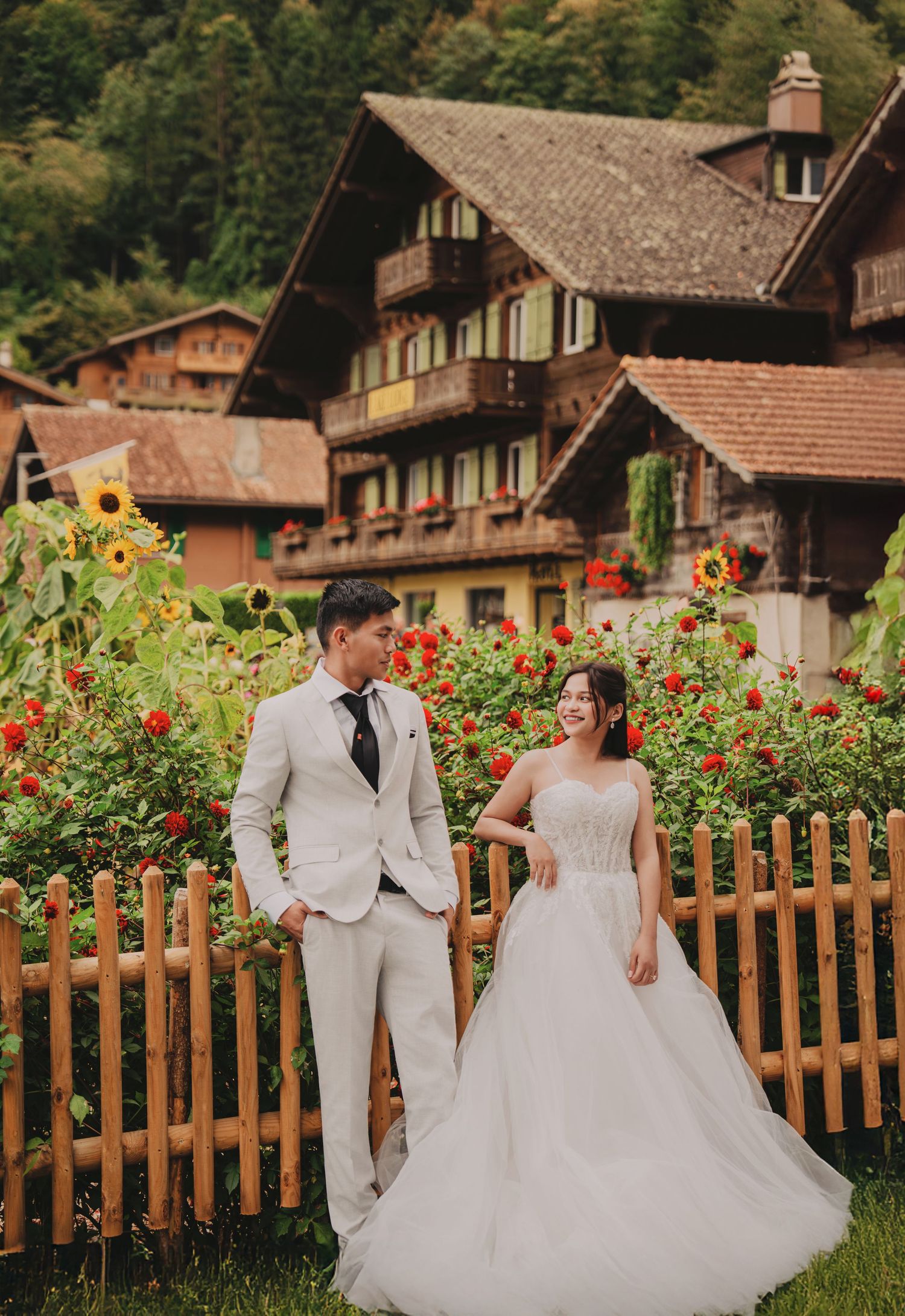 Wedding couple holding hands in garden with sunflowers and traditional Swiss chalet in lush green setting.