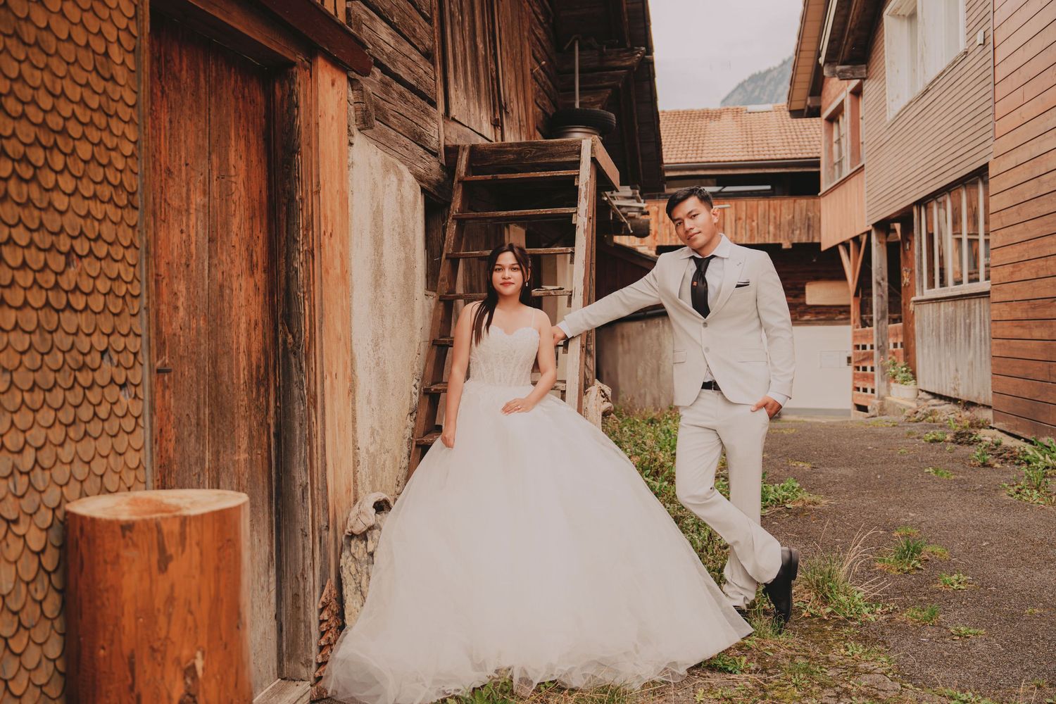 Bride in white ballgown and groom in ivory suit pose playfully beside a rustic wooden barn.
