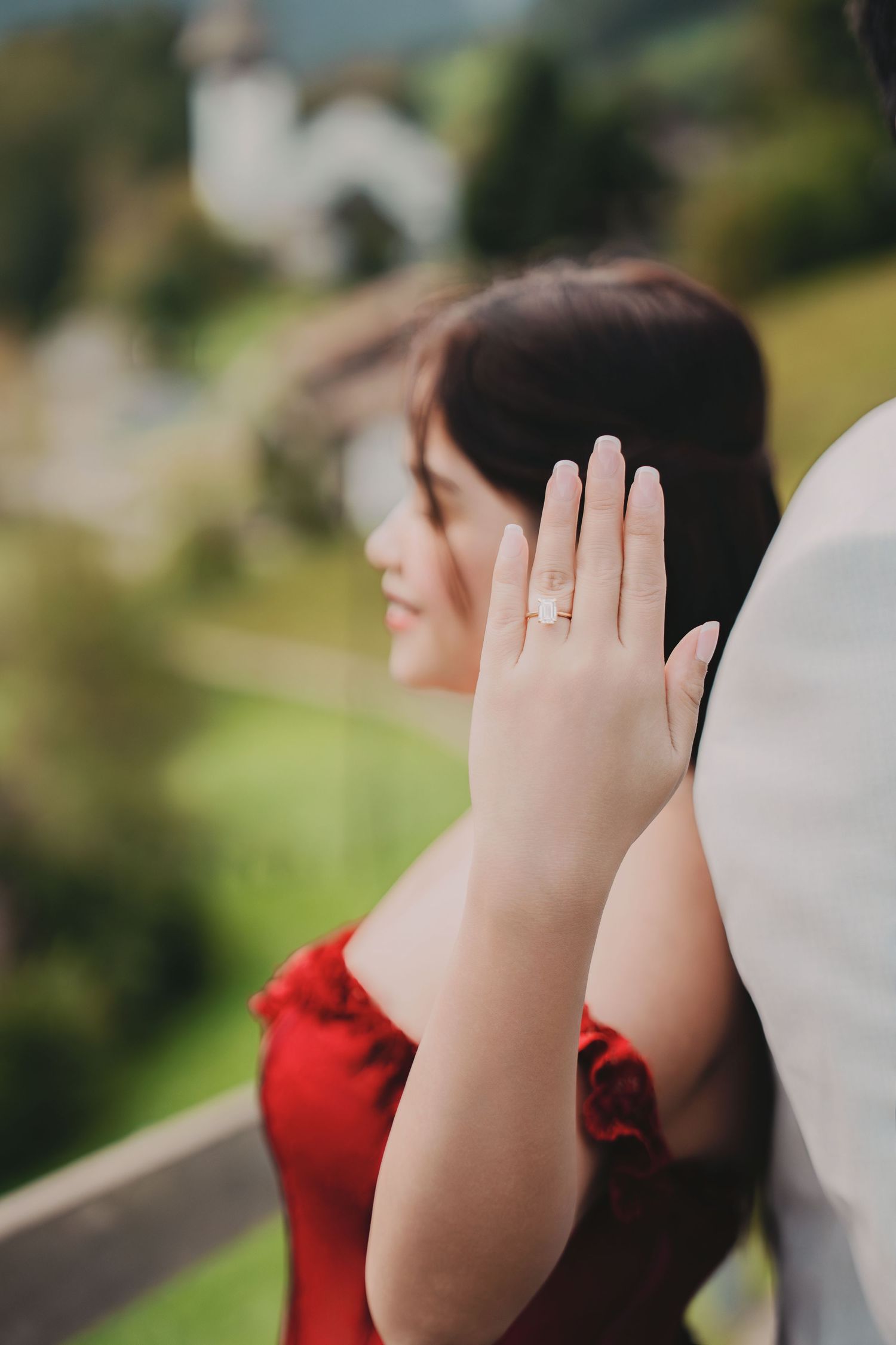 Bride in red off-shoulder gown displaying diamond engagement ring against scenic Swiss village backdrop.