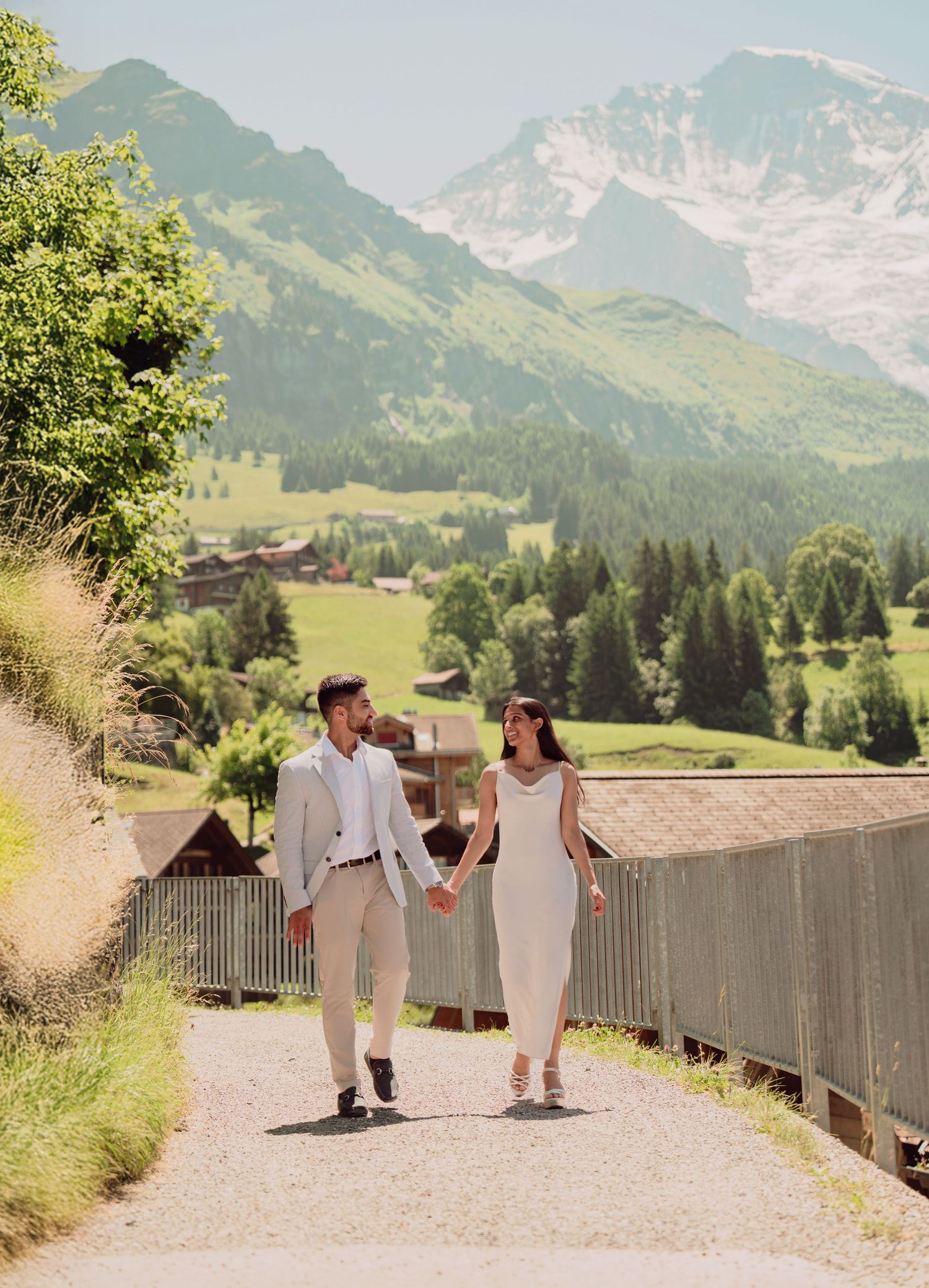 Stylish couple holding hands strolling along a scenic path with lush Swiss Alpine landscape backdrop.