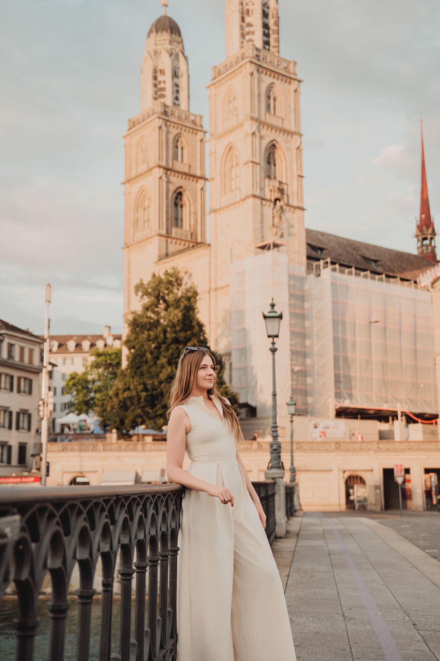Woman in white jumpsuit poses gracefully by iron fence with twin towers of Grossmunster Cathedral, Zurich.