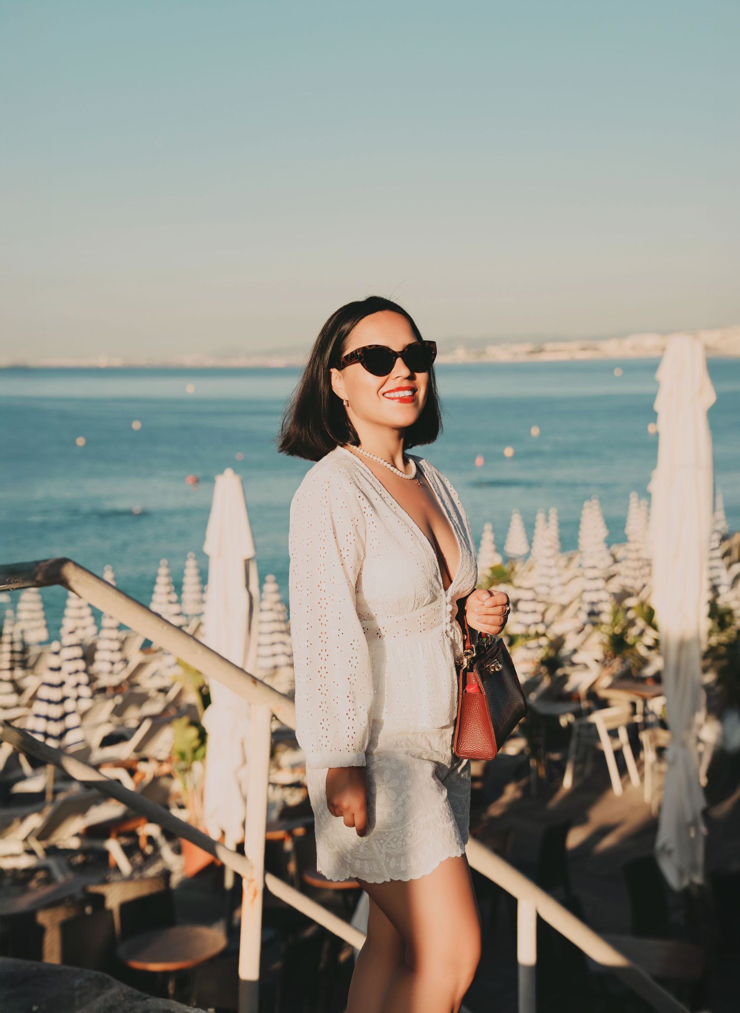 Stylish woman in white dress and sunglasses standing near seaside beach club.