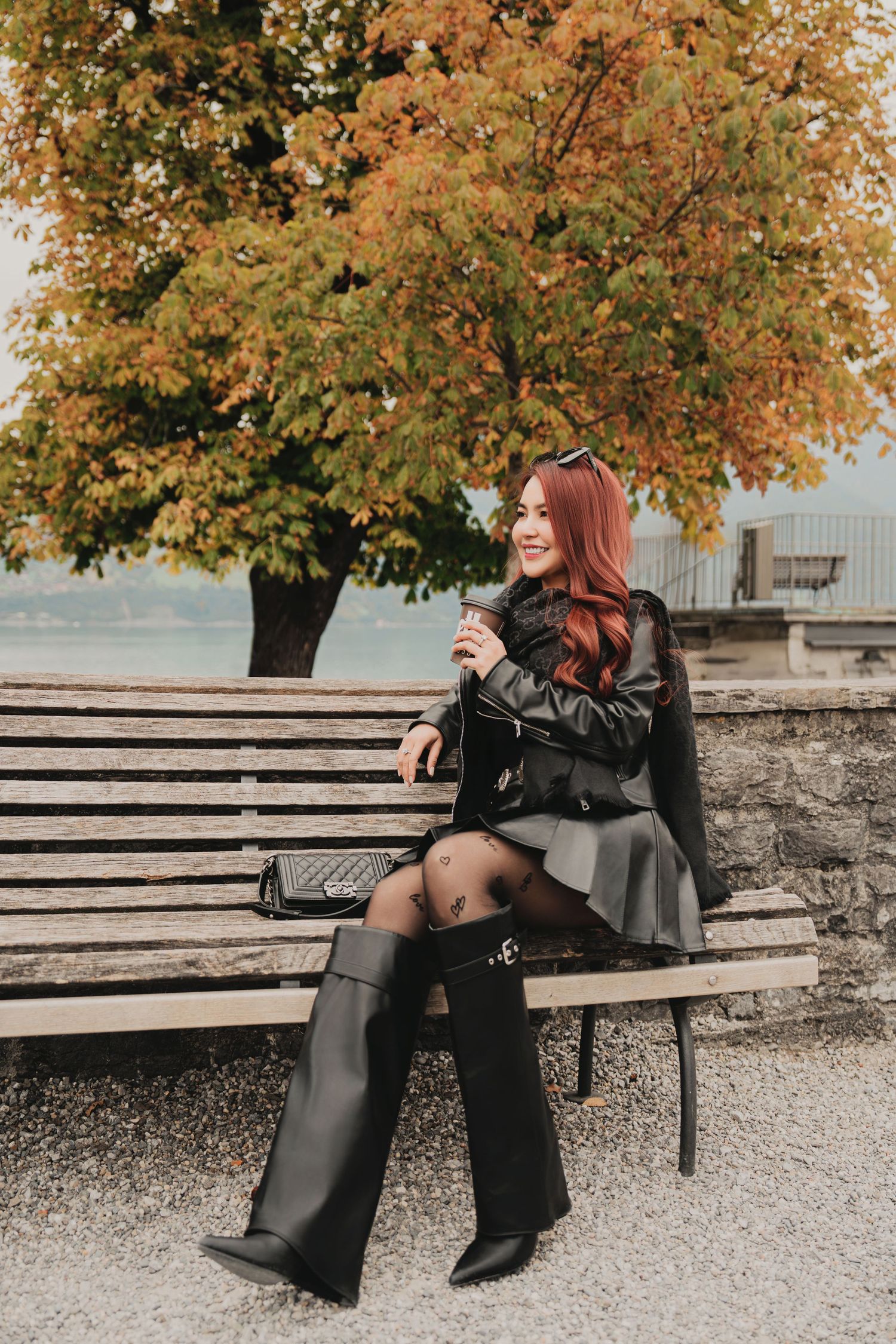 Red-haired woman in leather jacket, gray skirt, and tall black boots sitting on a park bench in autumn.