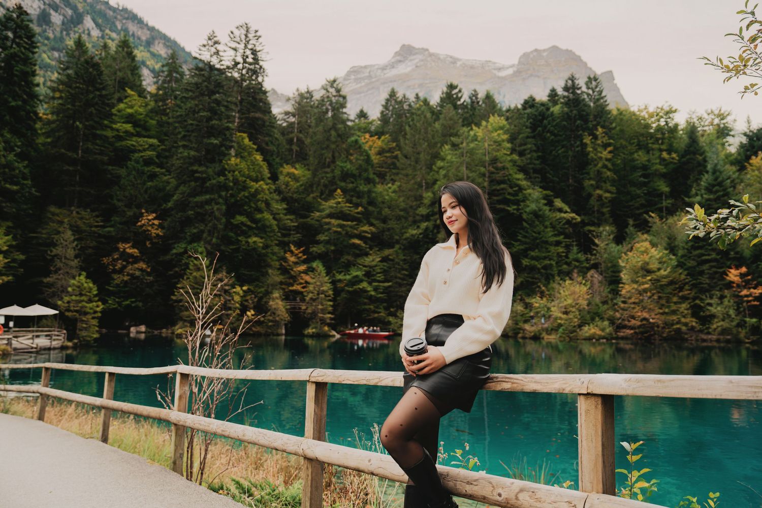 Woman in white sweater and black skirt leaning on wooden fence by turquoise alpine lake with mountain backdrop.