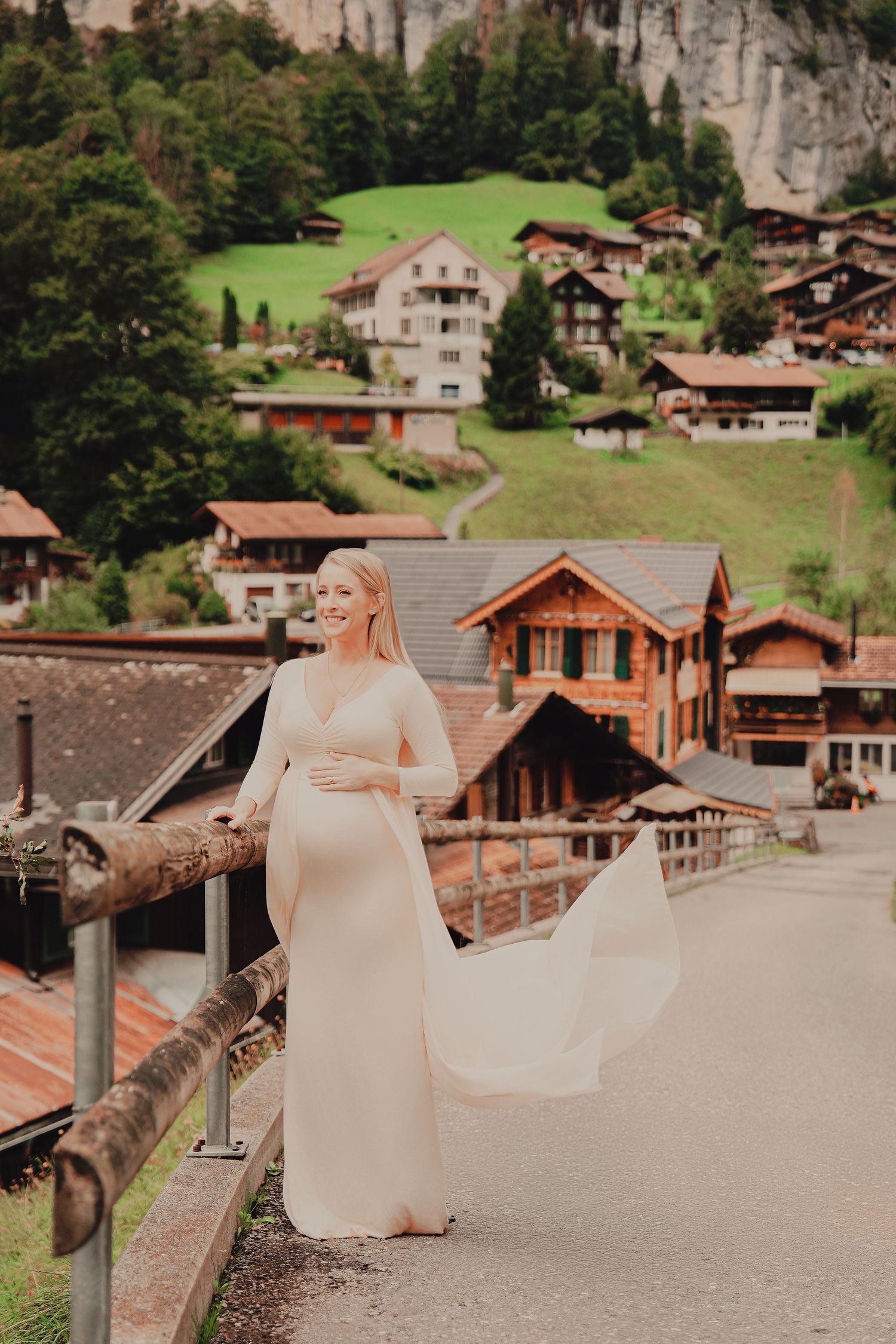 Smiling pregnant woman in flowing white gown stands on bridge with Swiss alpine village of Lauterbrunnen behind her.