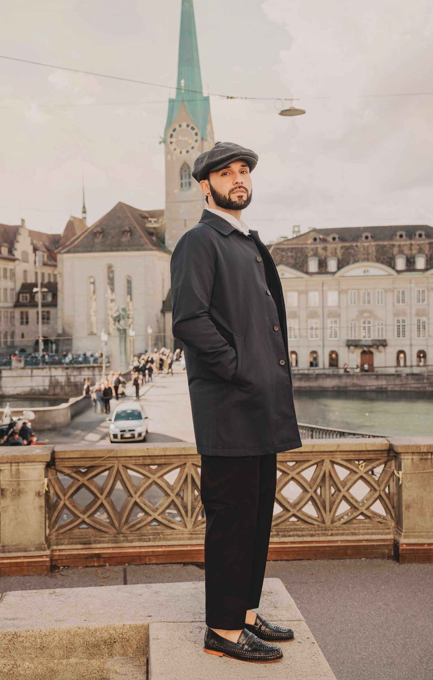 Fashionable bearded man in dark coat and flat cap stands on Zurich bridge with Fraumunster church behind.