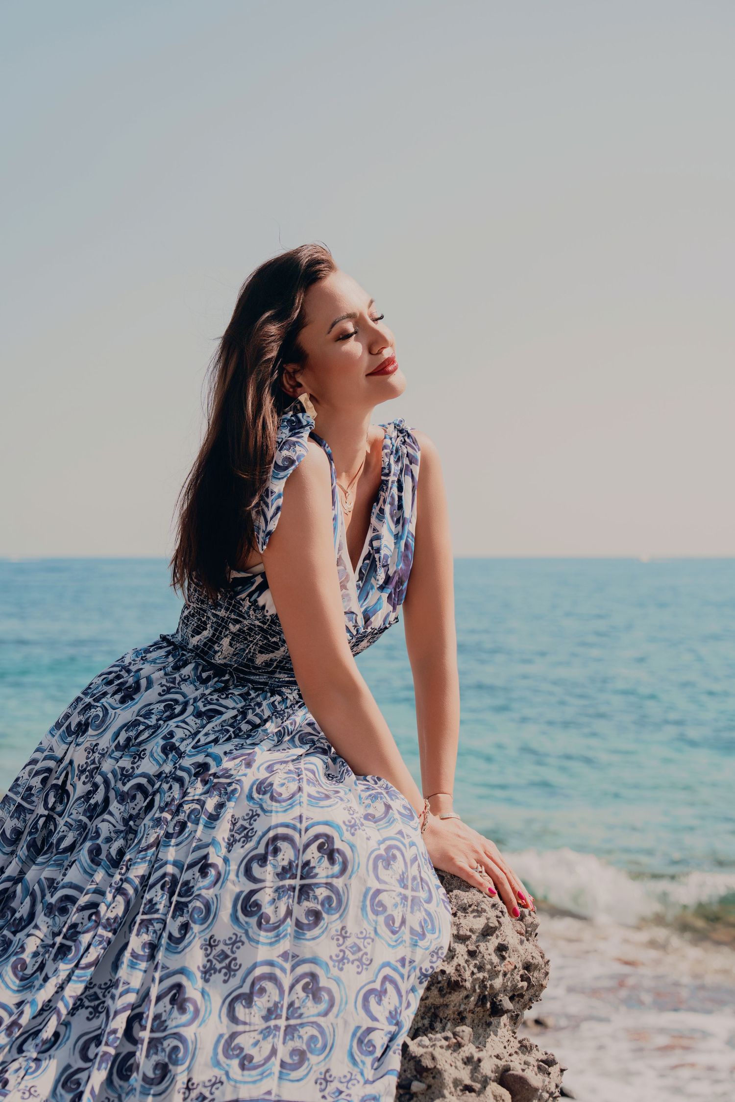 Beautiful woman in blue patterned sundress sitting on seaside rocks with eyes closed in sunlight.