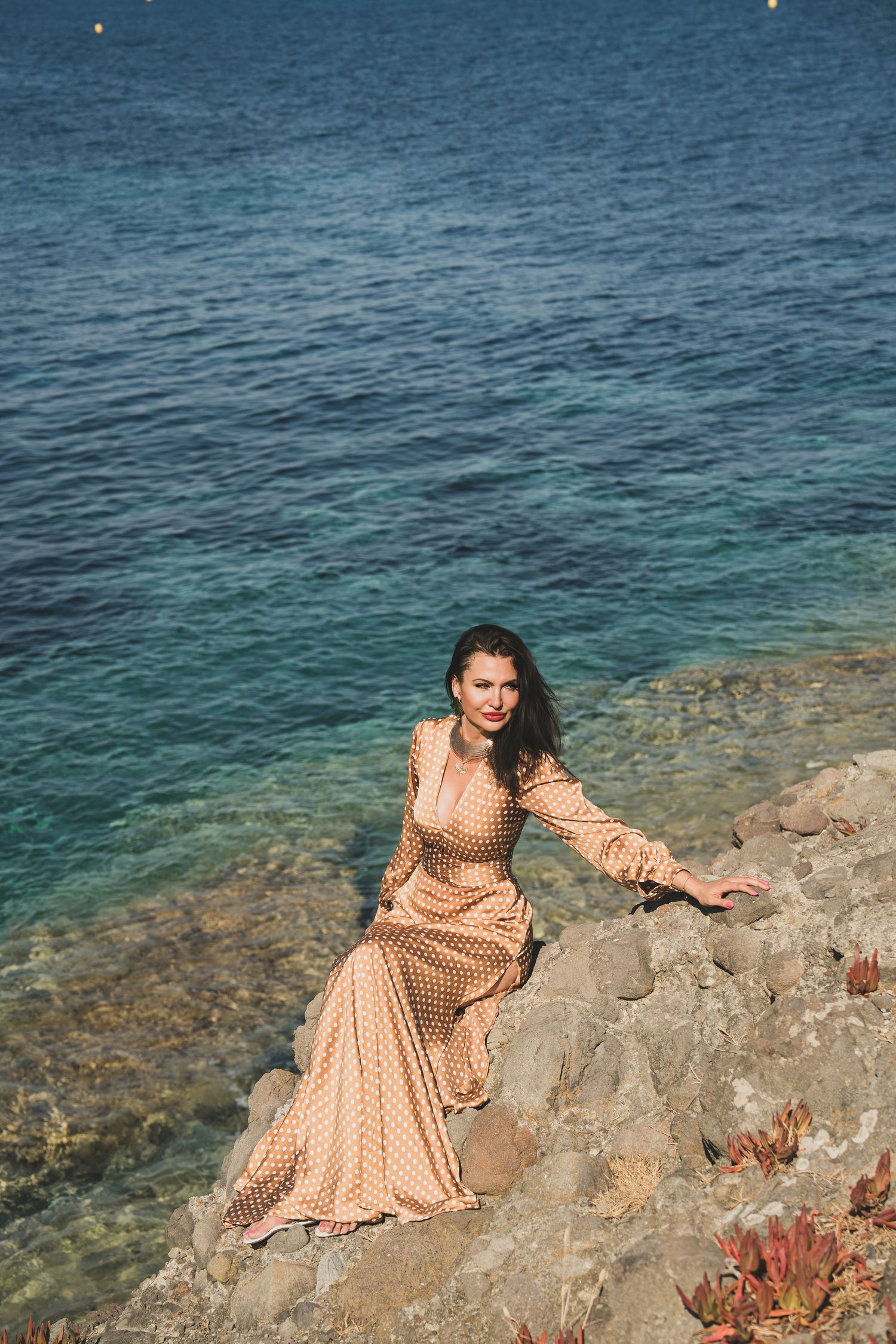 Beautiful woman in beige polka dot maxi dress posing on rocks by turquoise sea.
