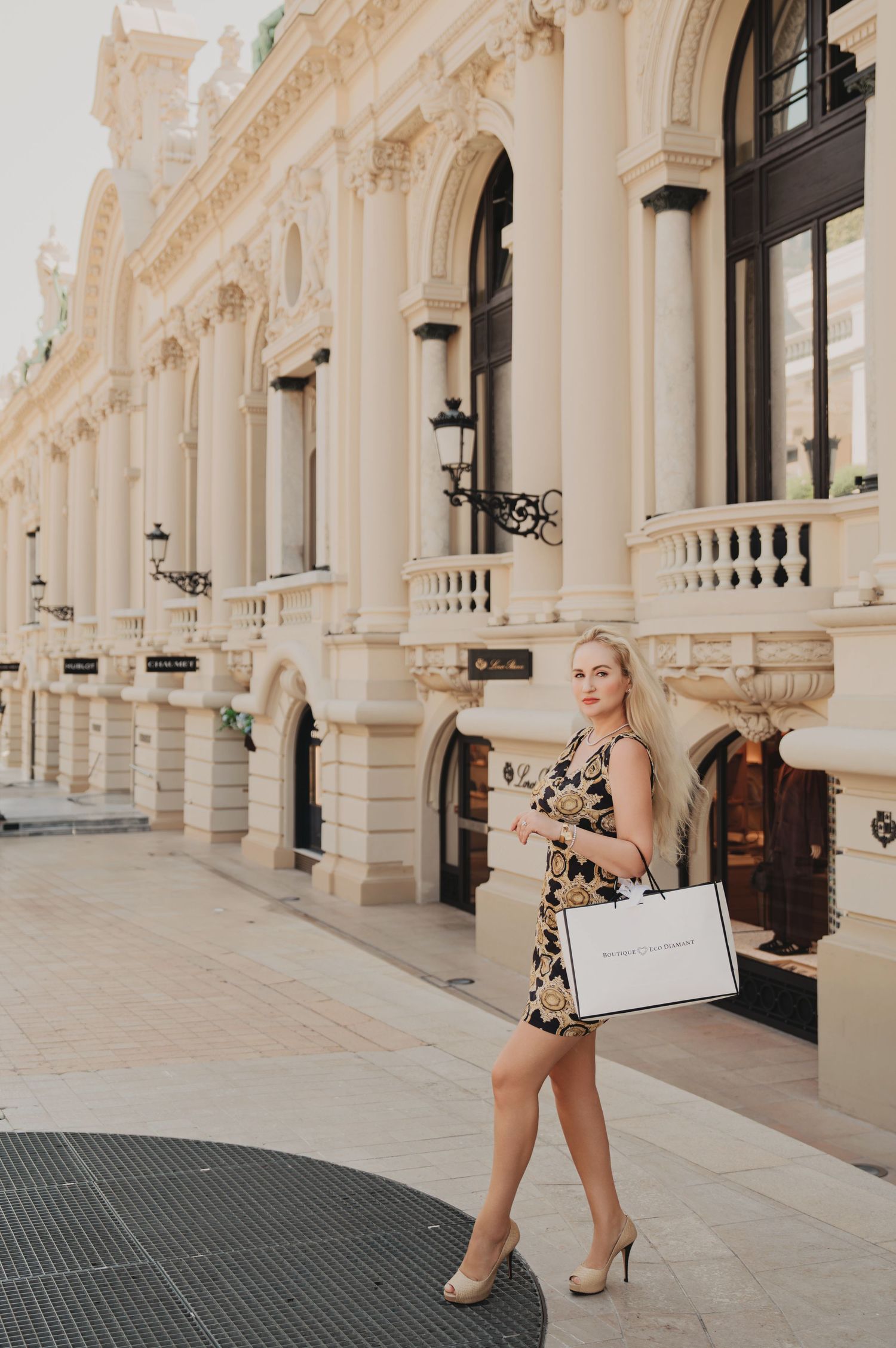 Blonde woman in black and gold print dress carrying luxury shopping bag near ornate building.
