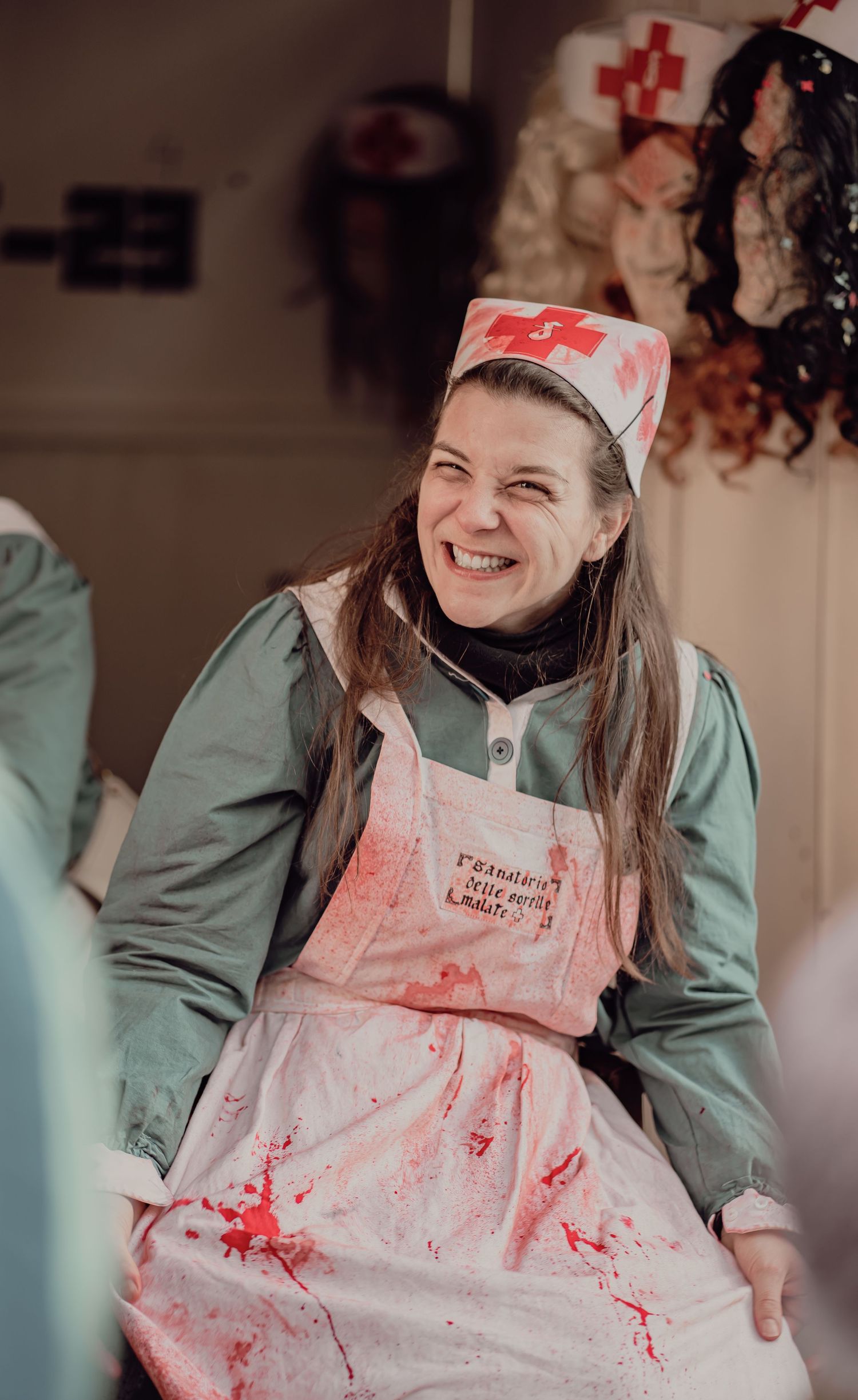 Woman in bloody nurse Halloween costume laughing joyfully at a festive indoor event.
