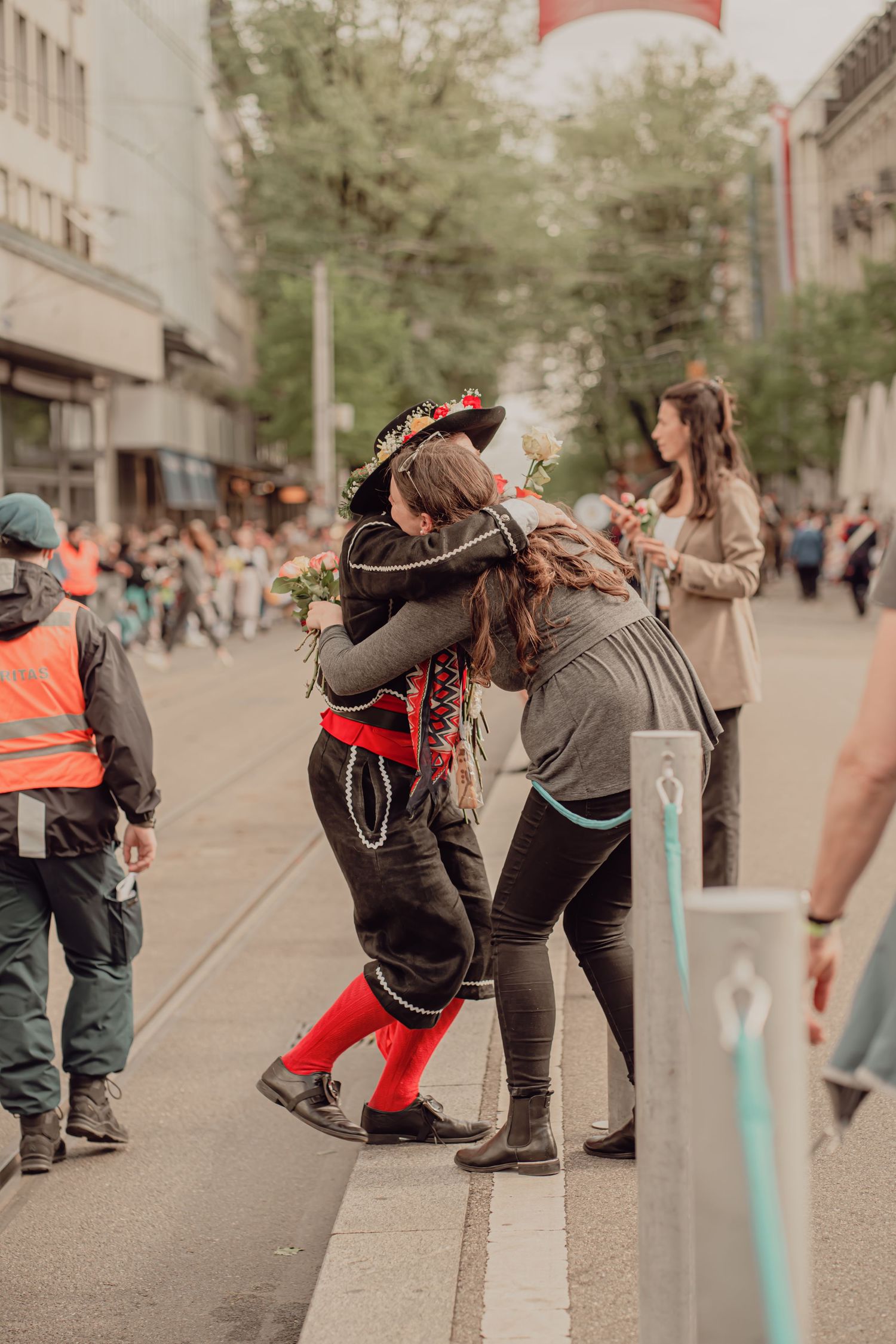 Two people in traditional folk costumes share a warm embrace on a busy parade street.