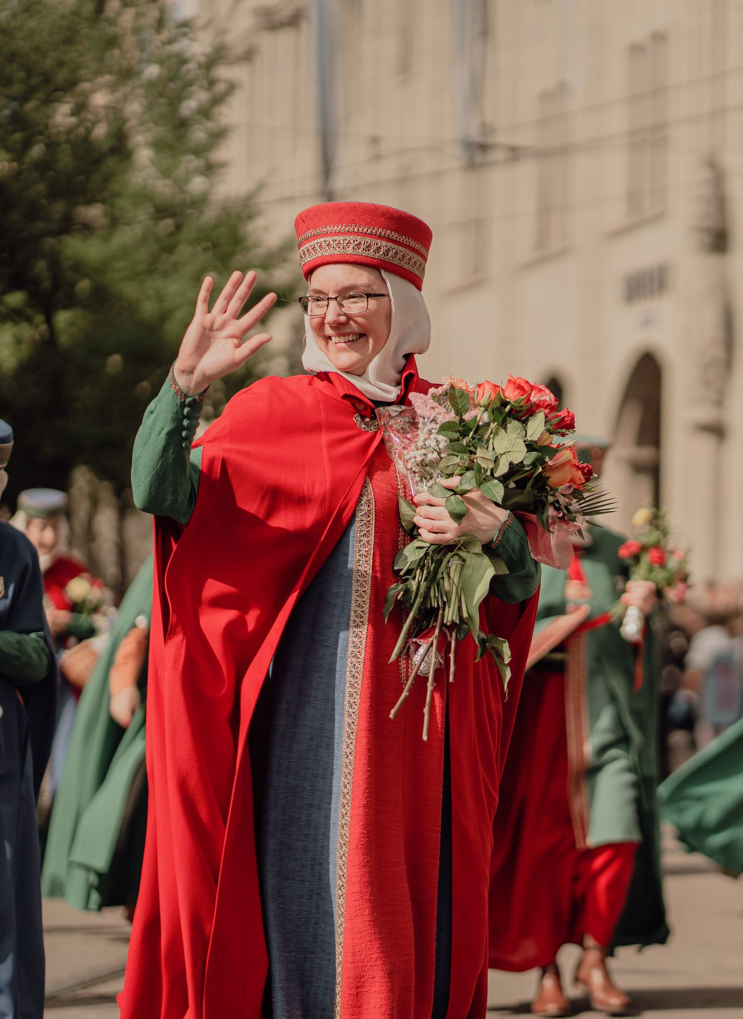 Woman in red medieval cape and hat waves while holding roses bouquet in a street parade.