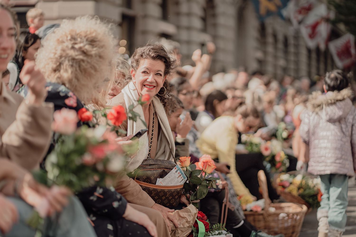 Smiling woman with flower baskets sits among cheerful spectators at a festive street parade.