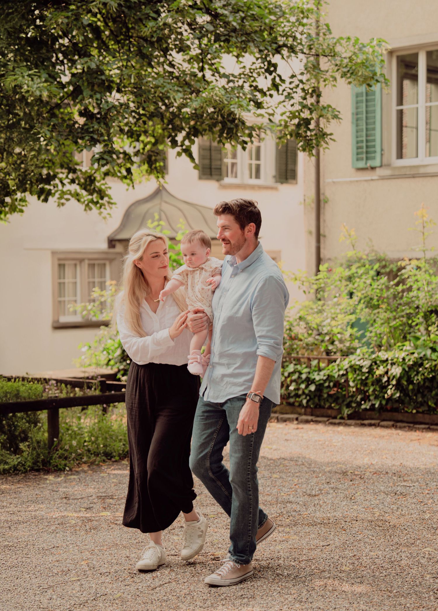 Happy young family strolling with baby in arms outside a historic European building with greenery.