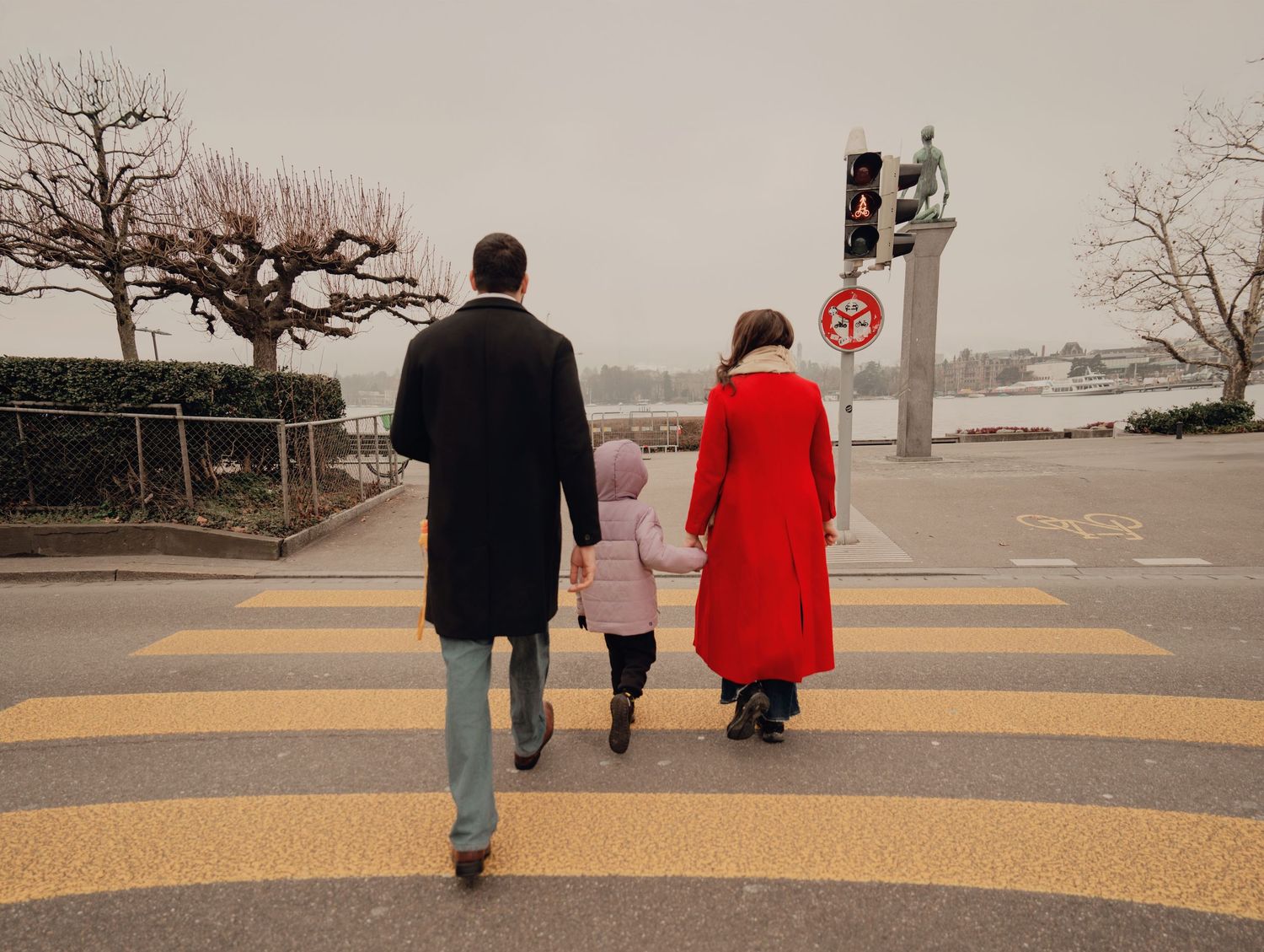 Family of three walks hand-in-hand across a yellow crosswalk on a foggy day near a lake.