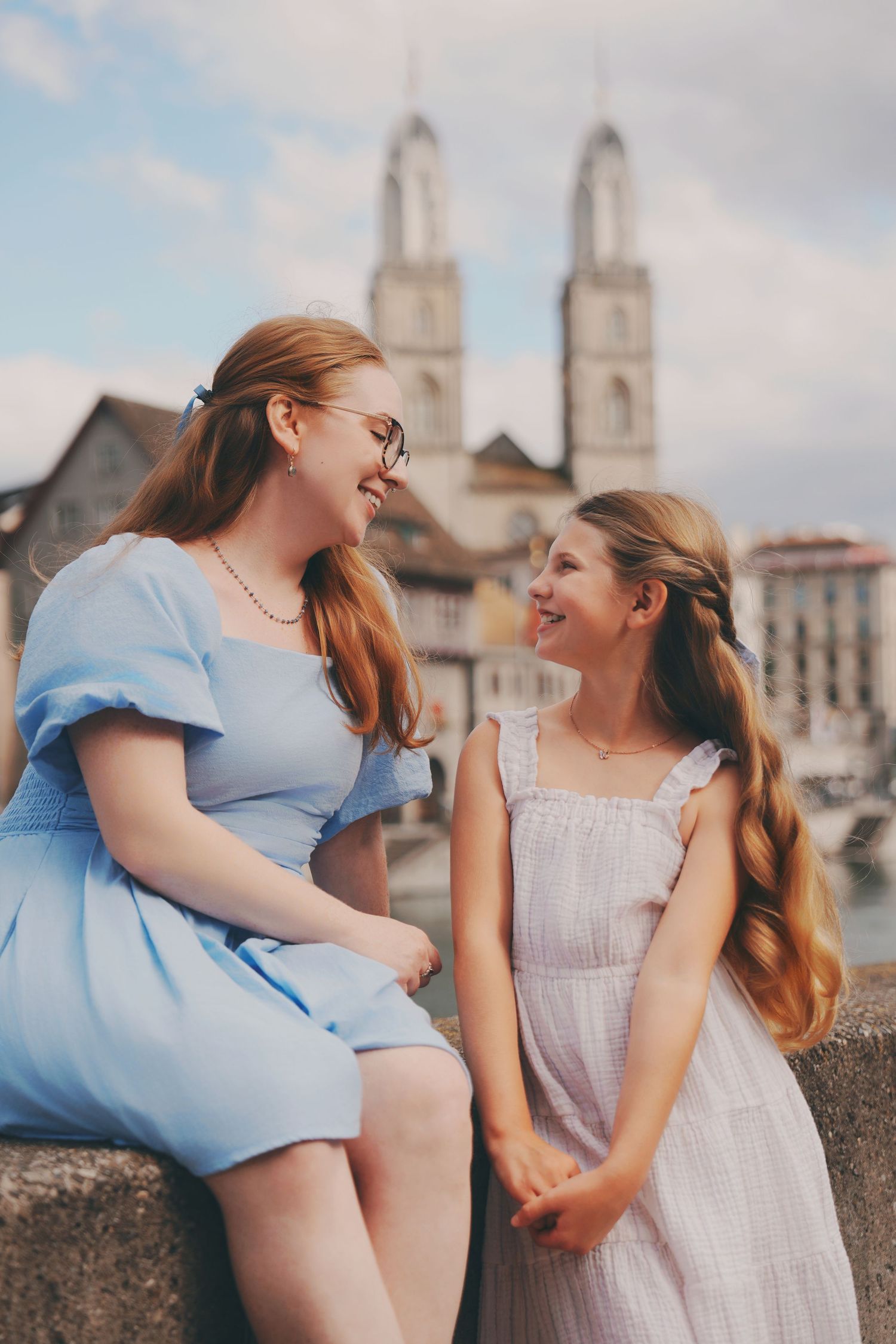 Mother and daughter smile at each other on stone bridge with Zurich cathedral in background.