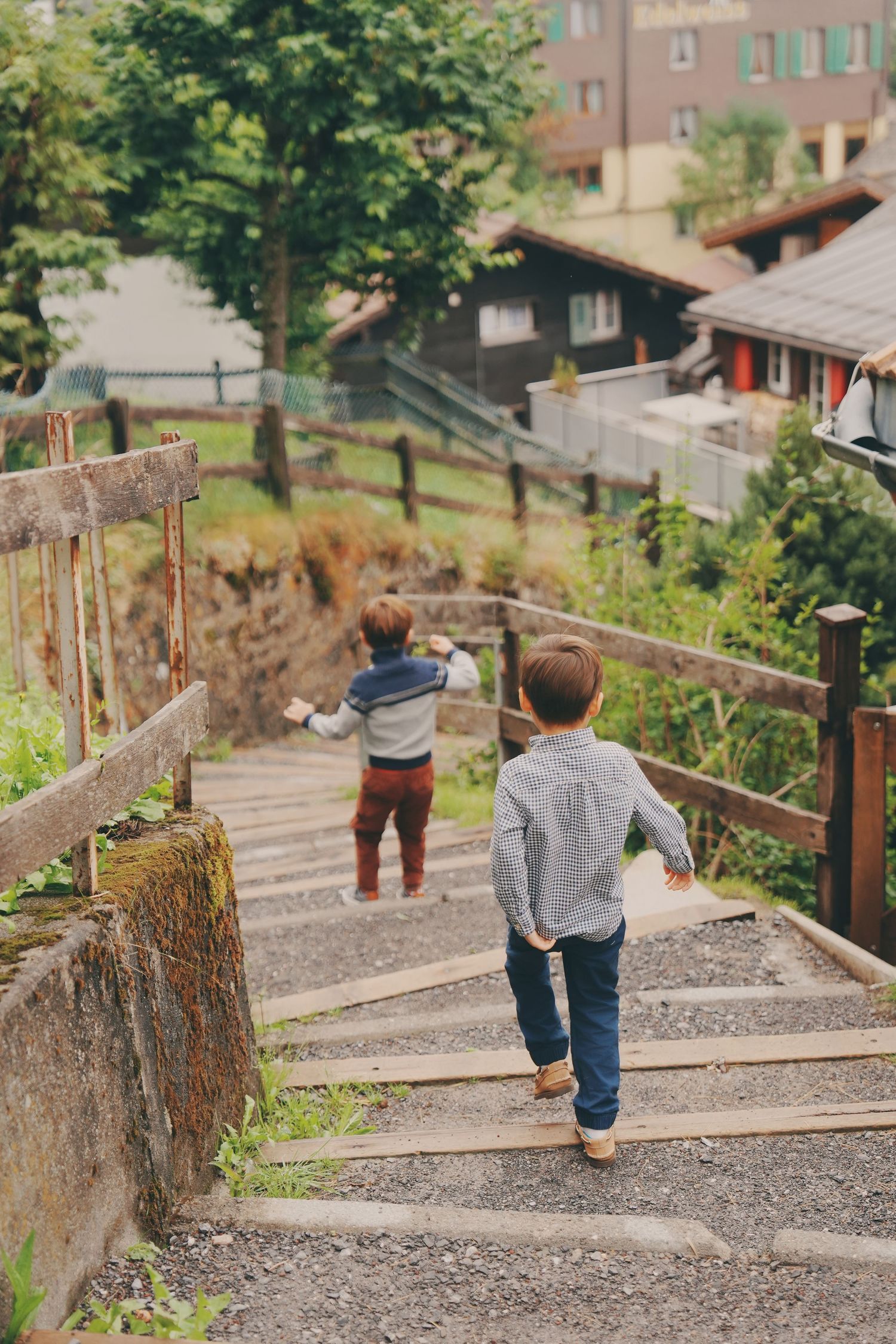 Two young boys climb wooden outdoor stairs in a charming alpine village surrounded by greenery.