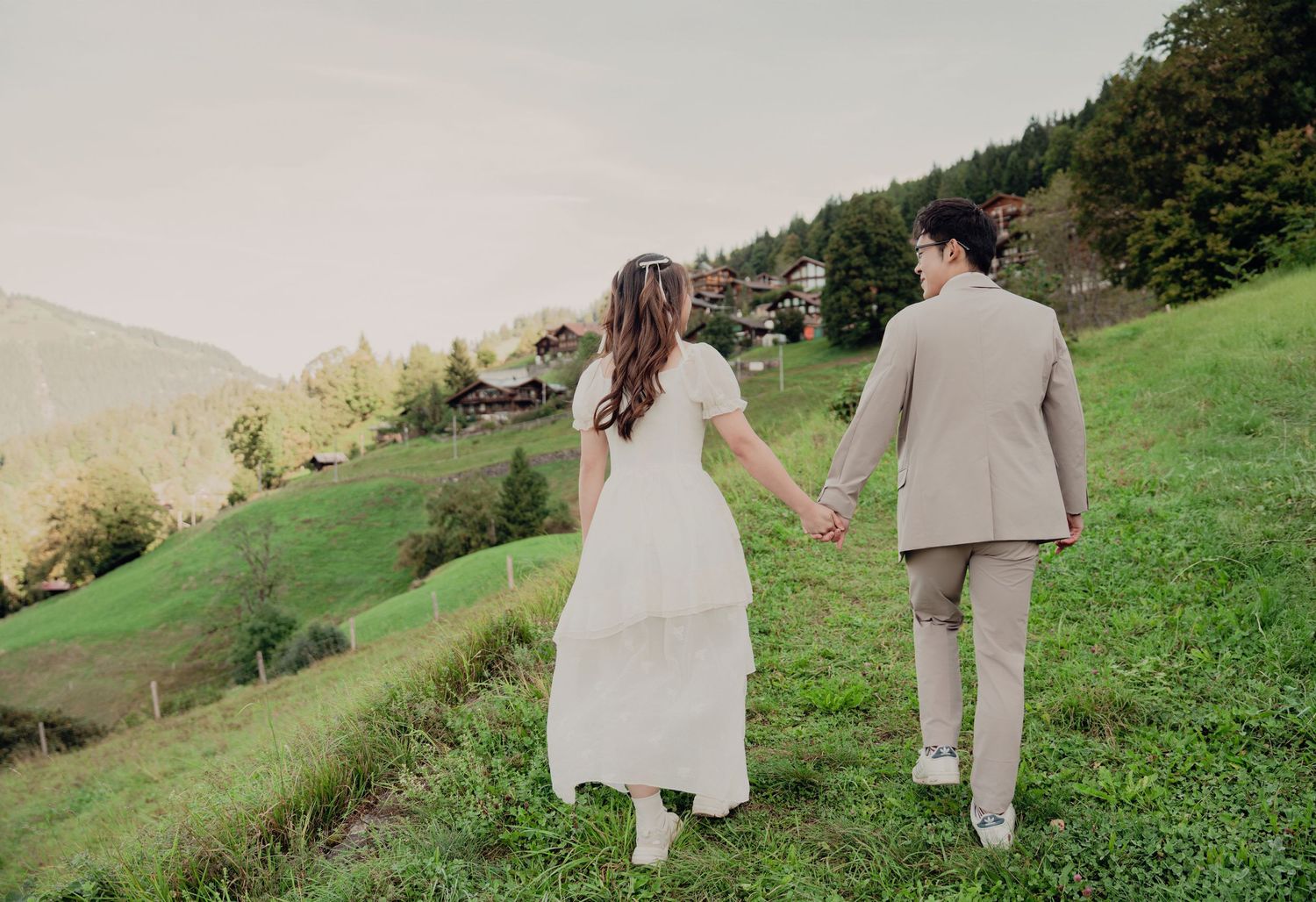 Couple in wedding attire holding hands walking on a lush green hillside with alpine village in background.