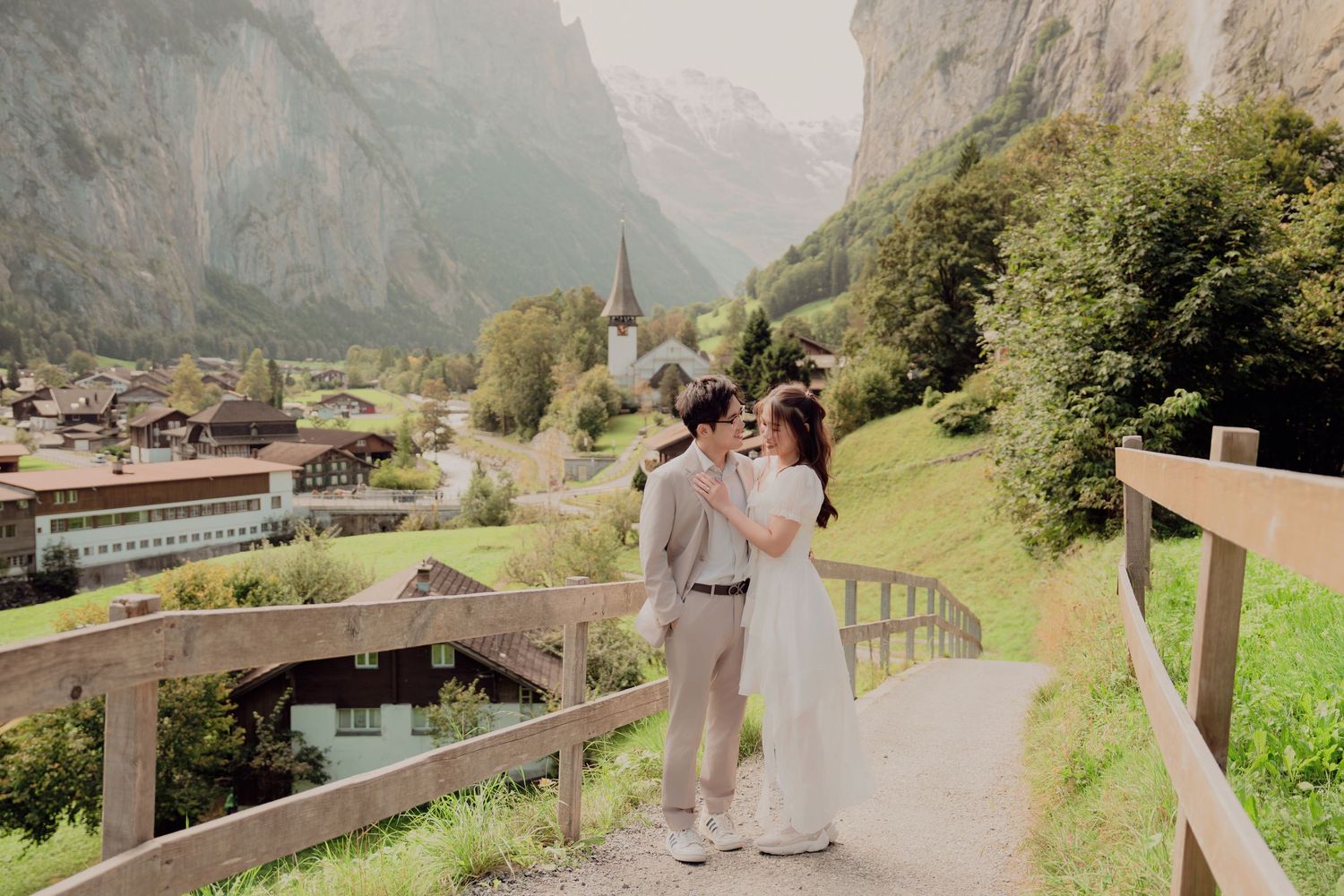 Couple embracing on wooden bridge path with scenic Swiss alpine village and dramatic mountain backdrop.