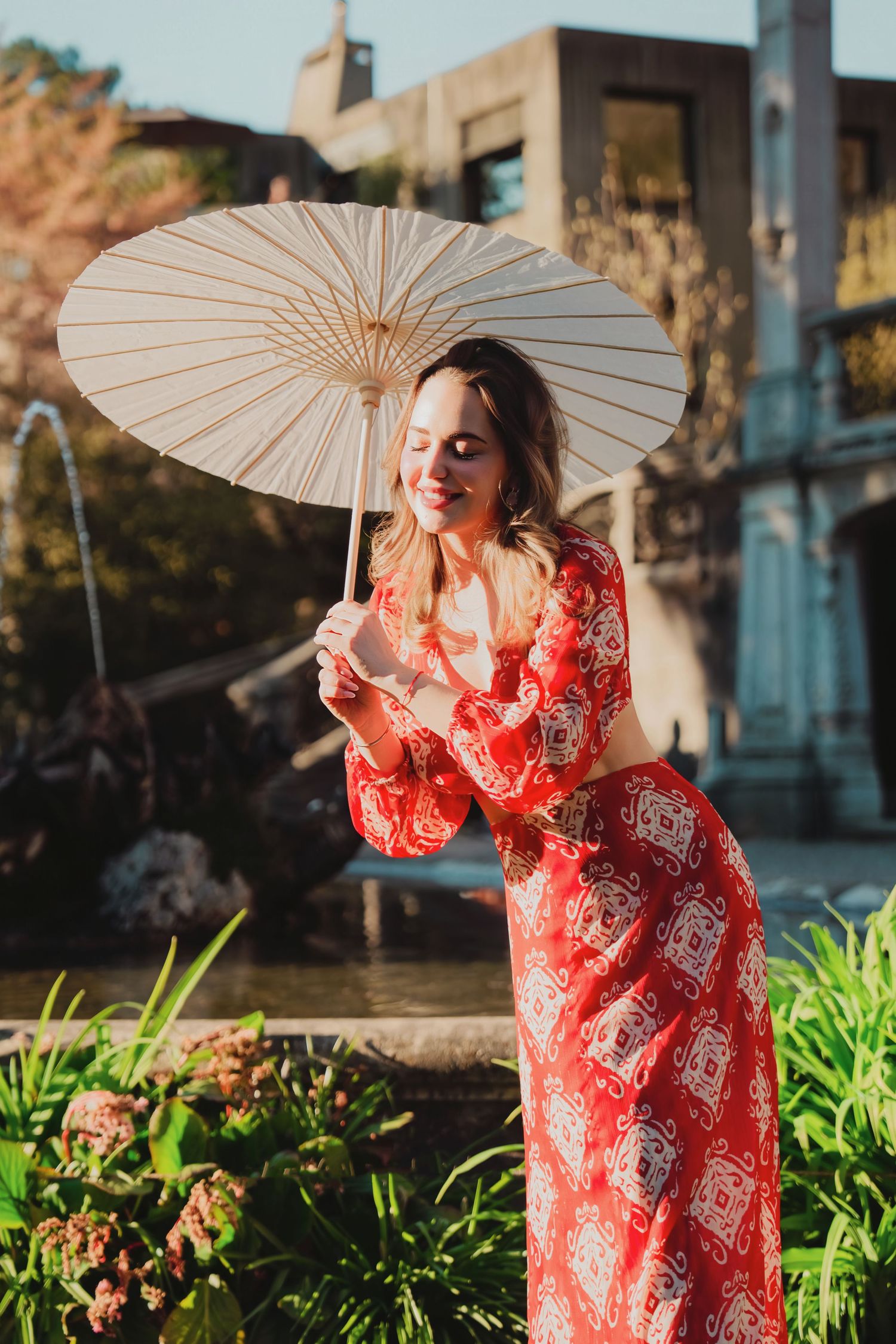 Woman in red patterned dress holding white paper parasol near garden fountain in warm sunlight.