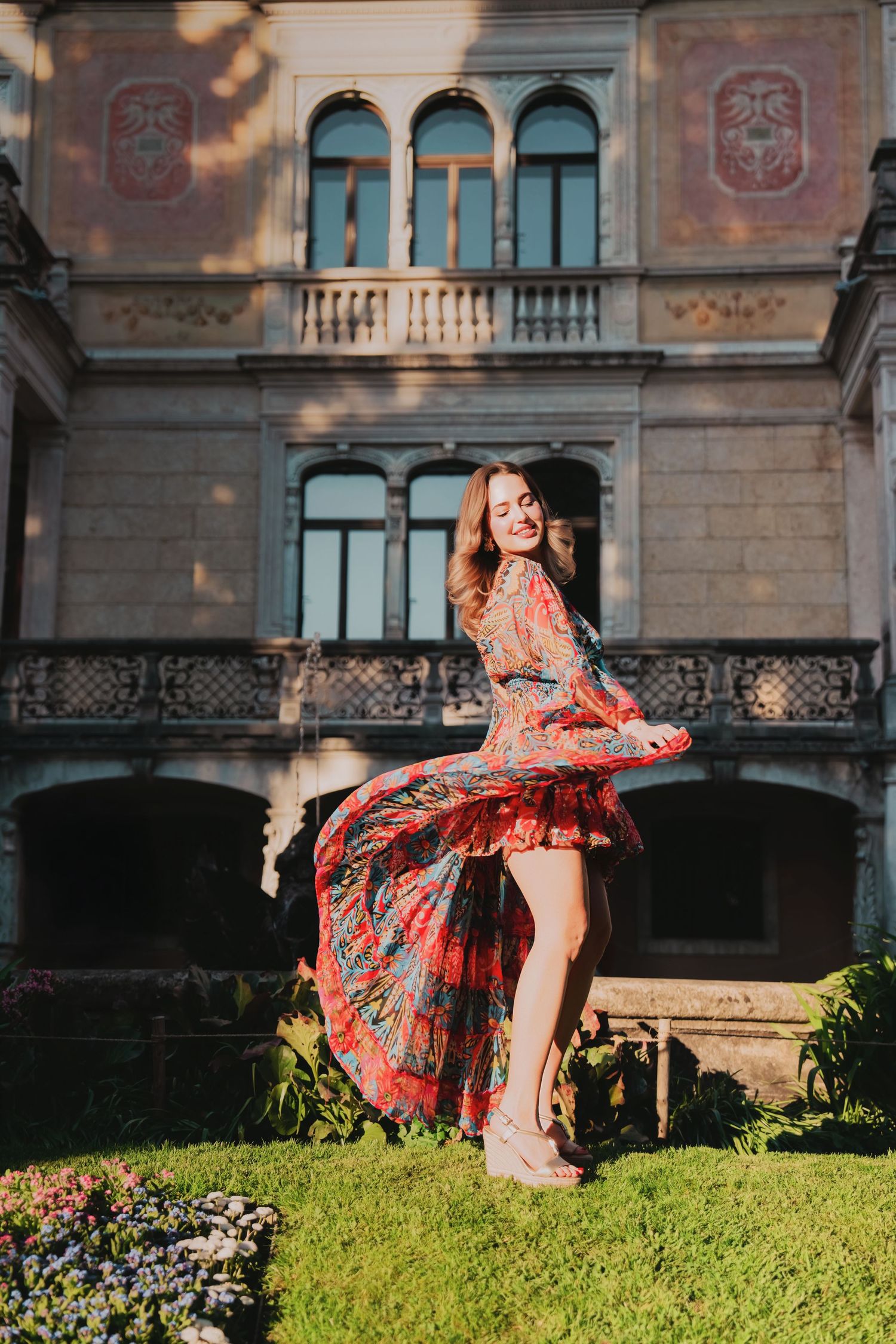 Woman twirling in colorful floral dress on green lawn before ornate historic European building.