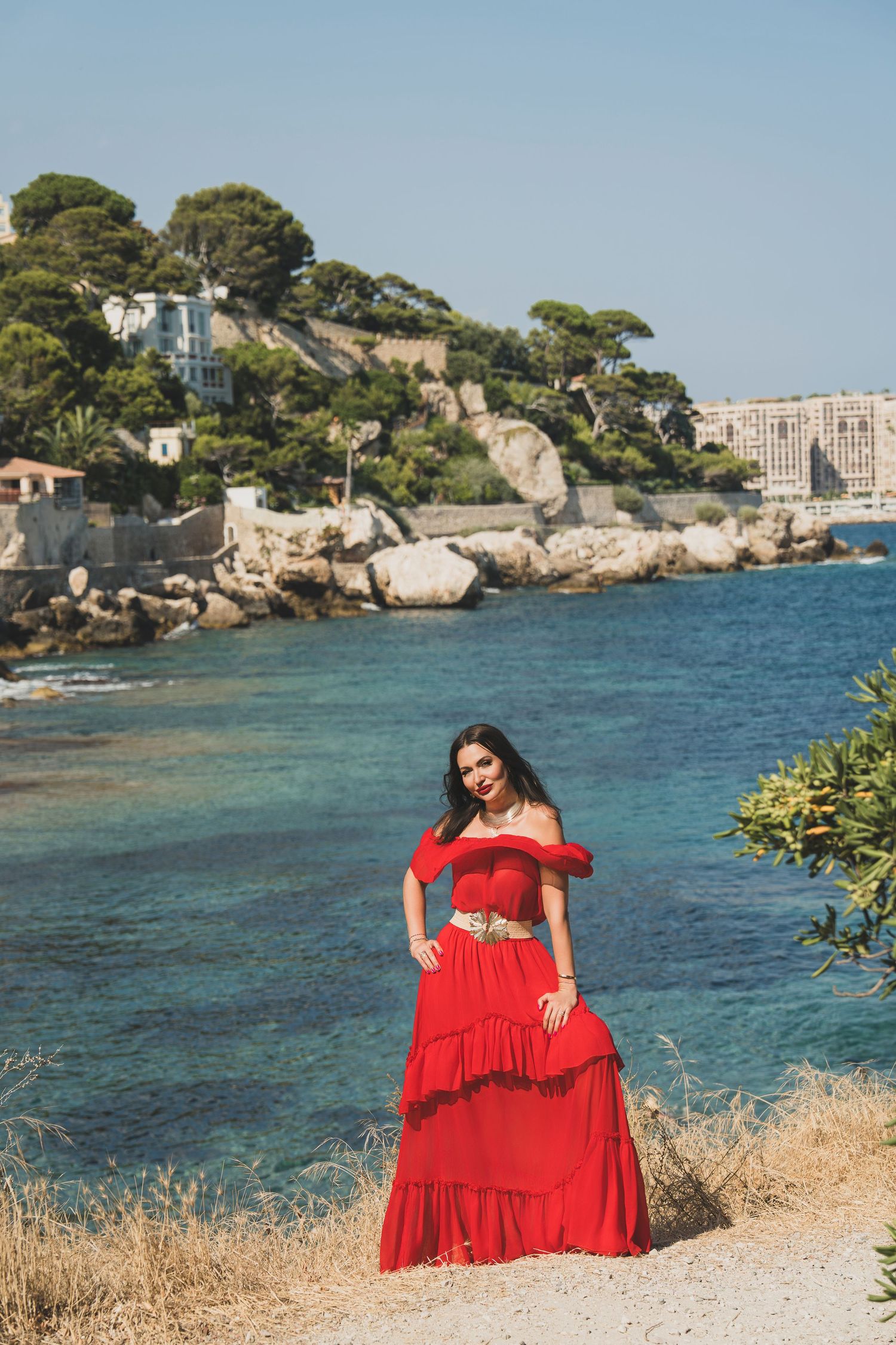 Brunette woman in red off-shoulder tiered maxi dress posing by turquoise coastal waters.