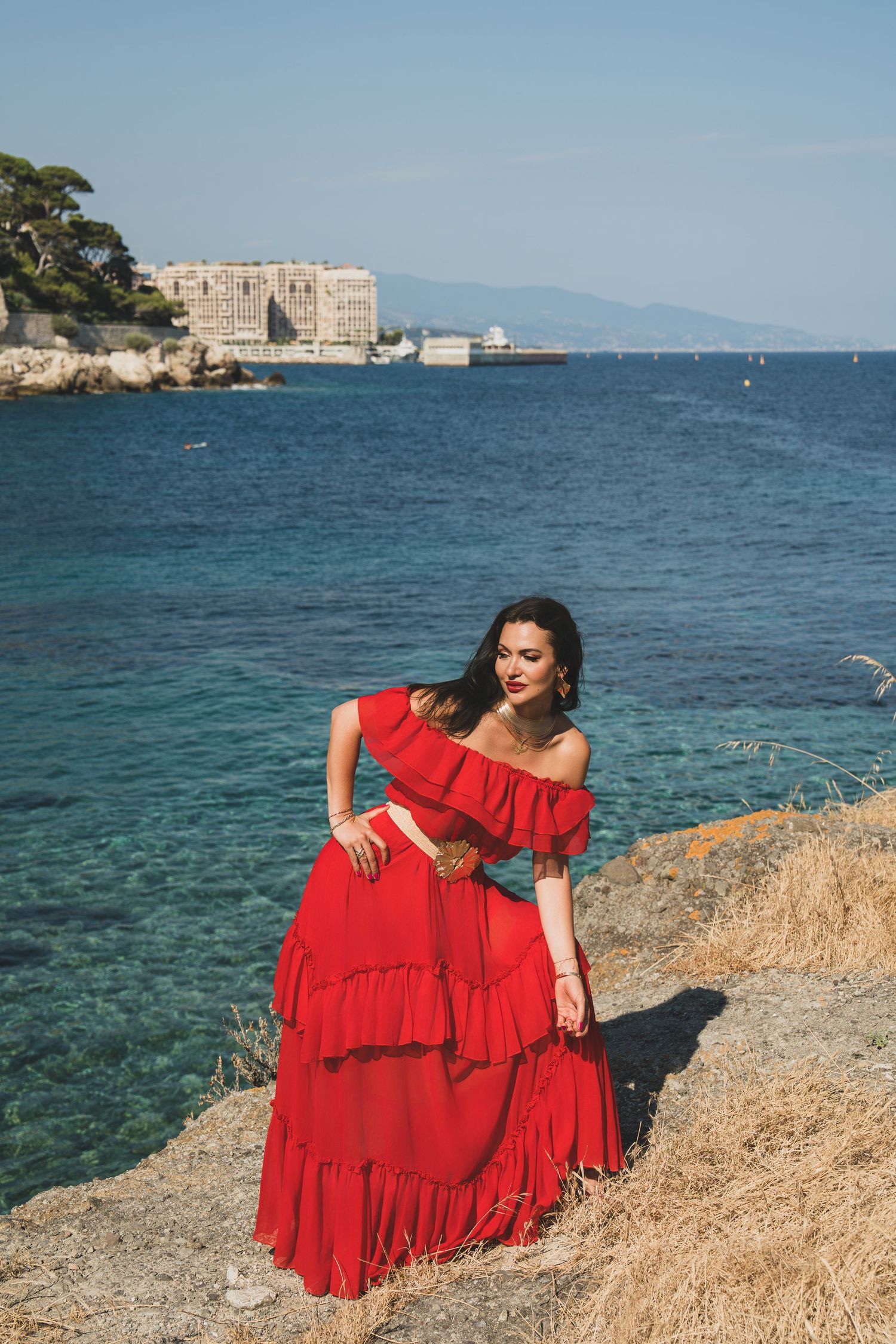Brunette woman in red ruffled off-shoulder maxi dress posing on rocky Mediterranean coastal landscape.