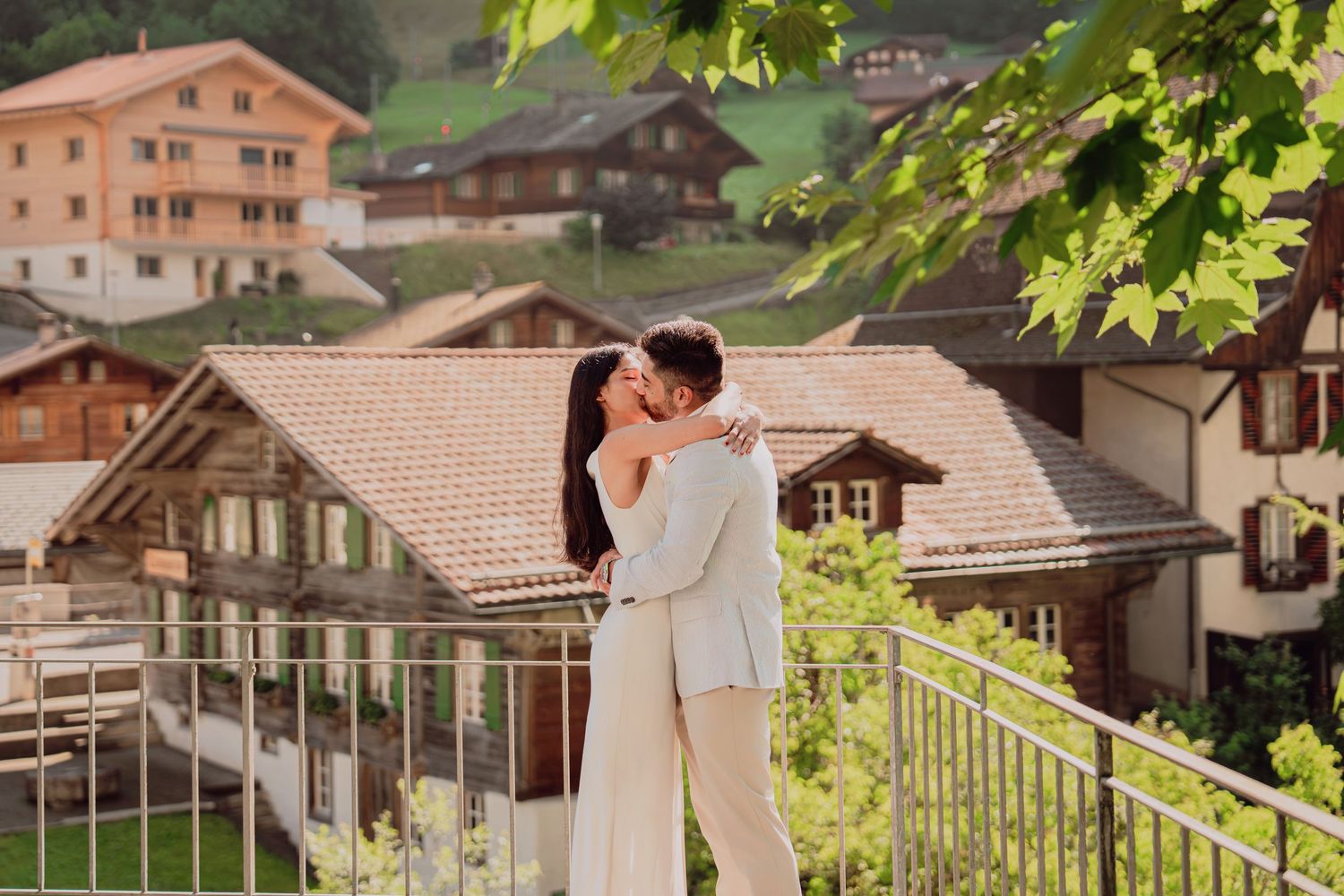 Couple sharing a kiss on a balcony with charming Swiss village rooftops behind them.