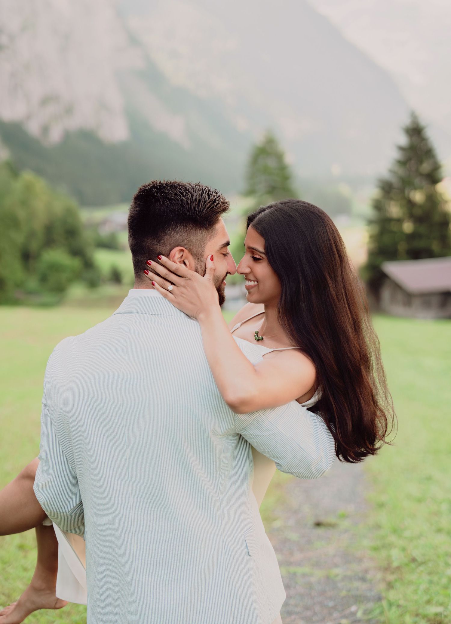 Happy couple embracing nose-to-nose in a Swiss alpine landscape with mountains and trees behind.