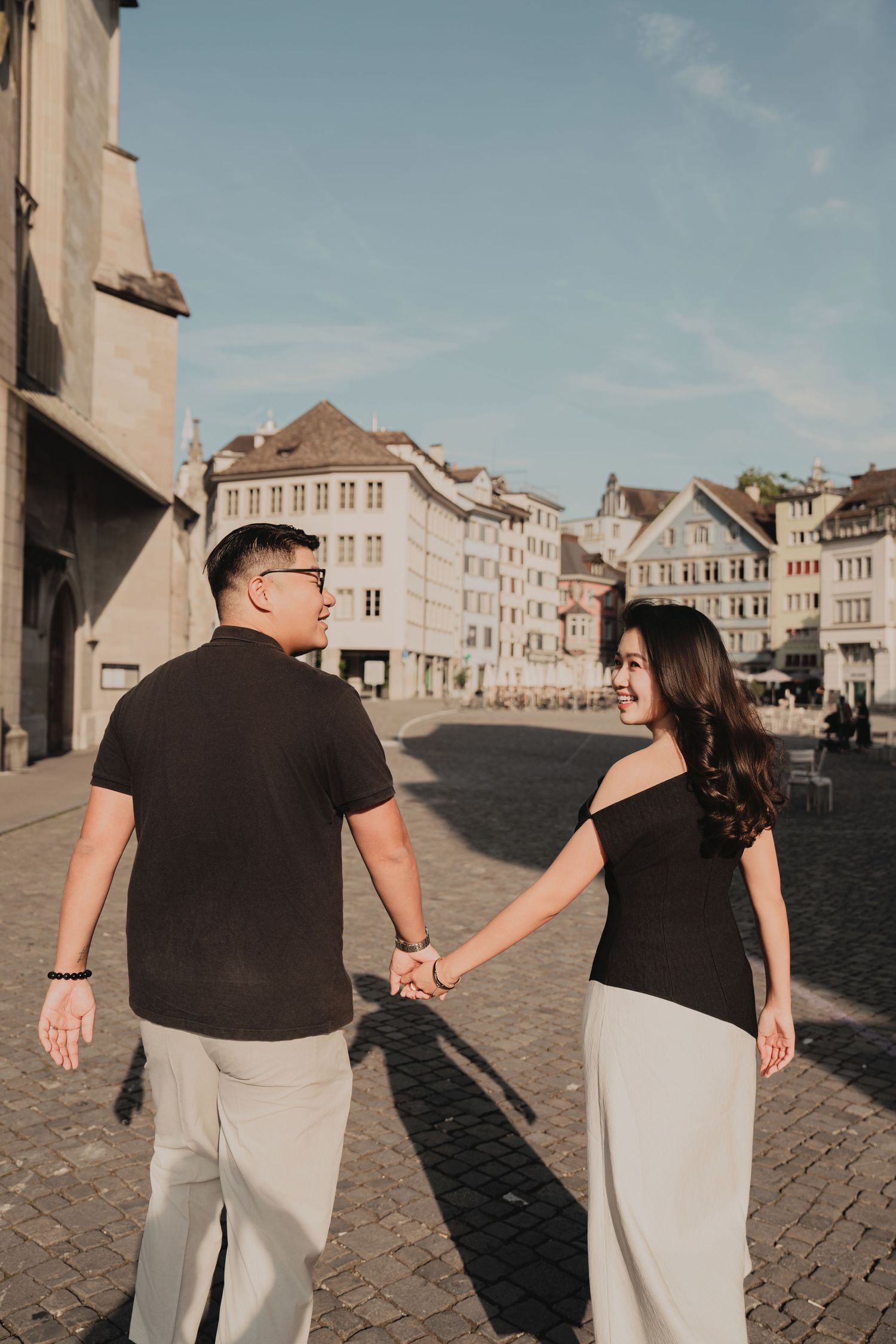 Young couple holding hands on a sunny cobblestone plaza, woman smiling back at camera in a European city.