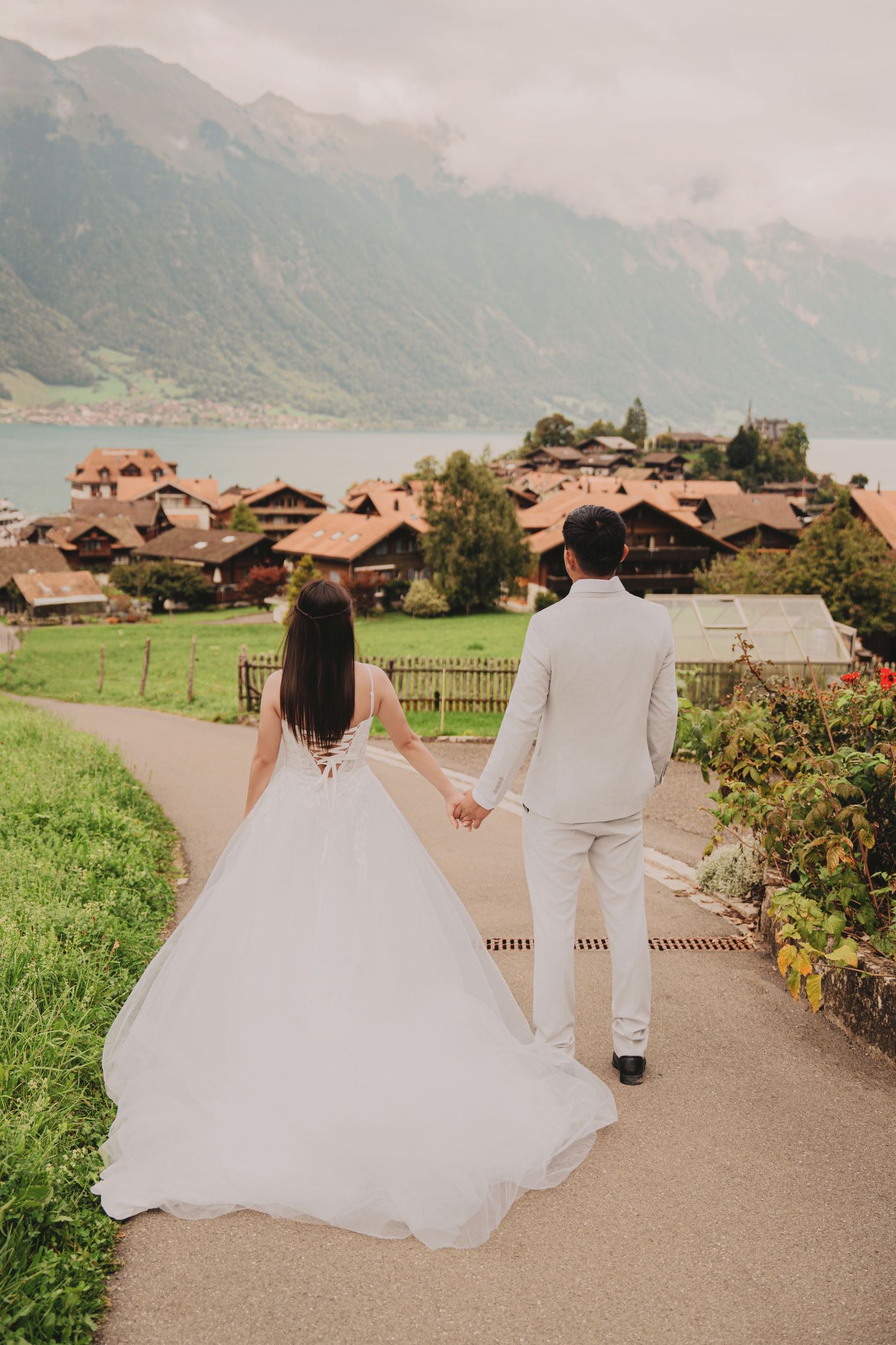 Wedding couple holding hands on path overlooking charming Swiss village with turquoise lake and mountains.