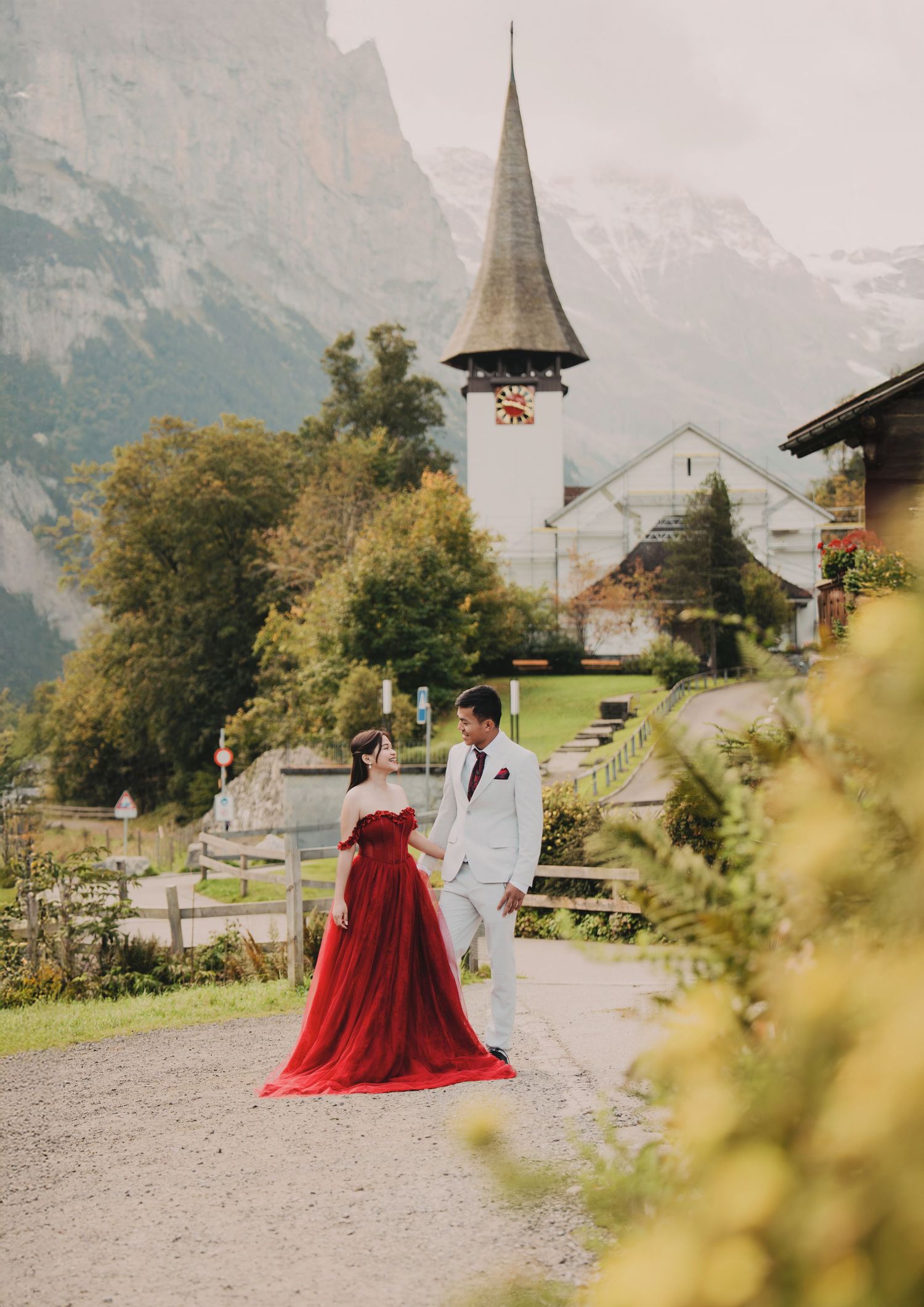 Elegant couple in red gown and white suit walking near Swiss alpine church with mountains.