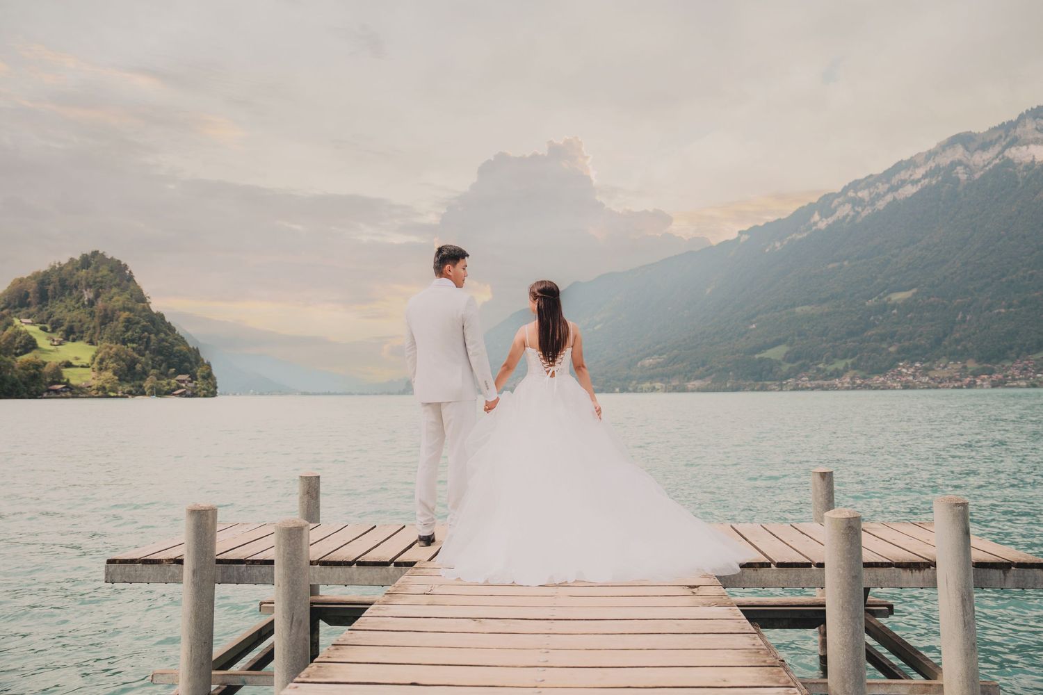 Bride and groom hold hands on a wooden pier gazing at a breathtaking alpine lake and mountain landscape.
