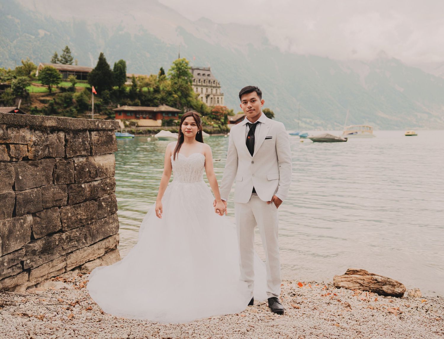 Elegant wedding couple holding hands on a rocky alpine lakeside with a historic mansion and mountains behind.