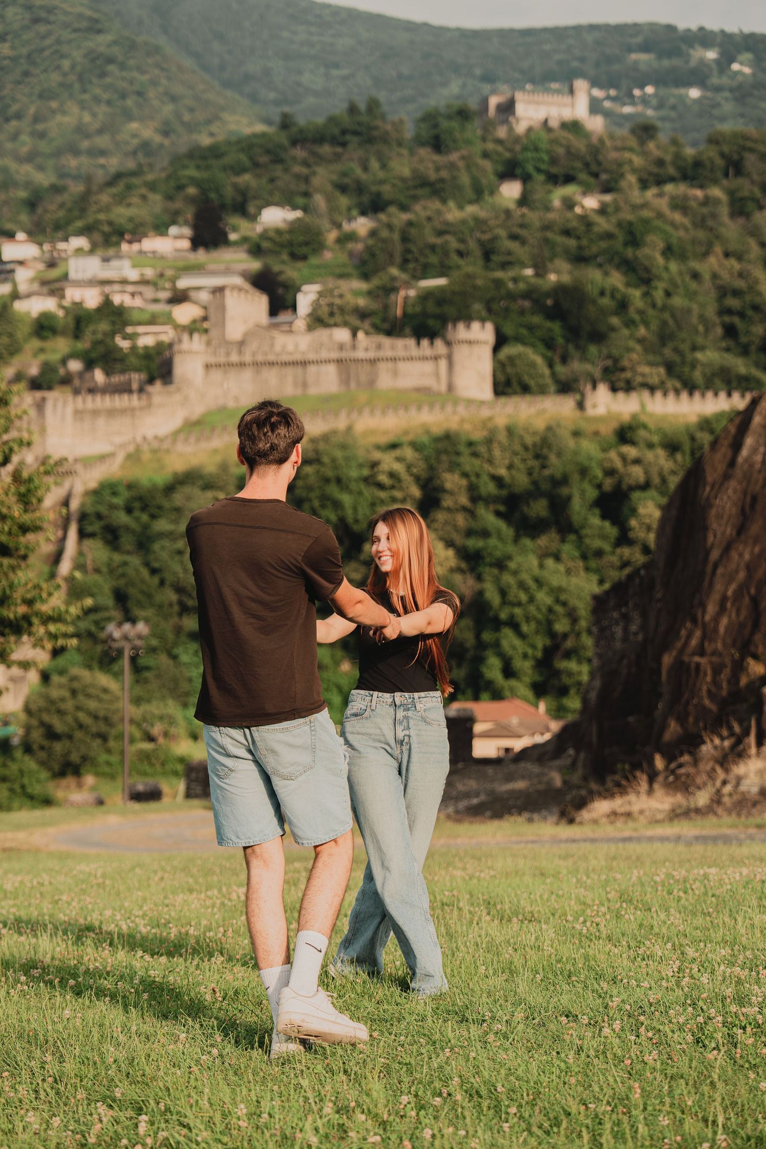 Couple dancing together on green hillside with medieval castle walls in background.