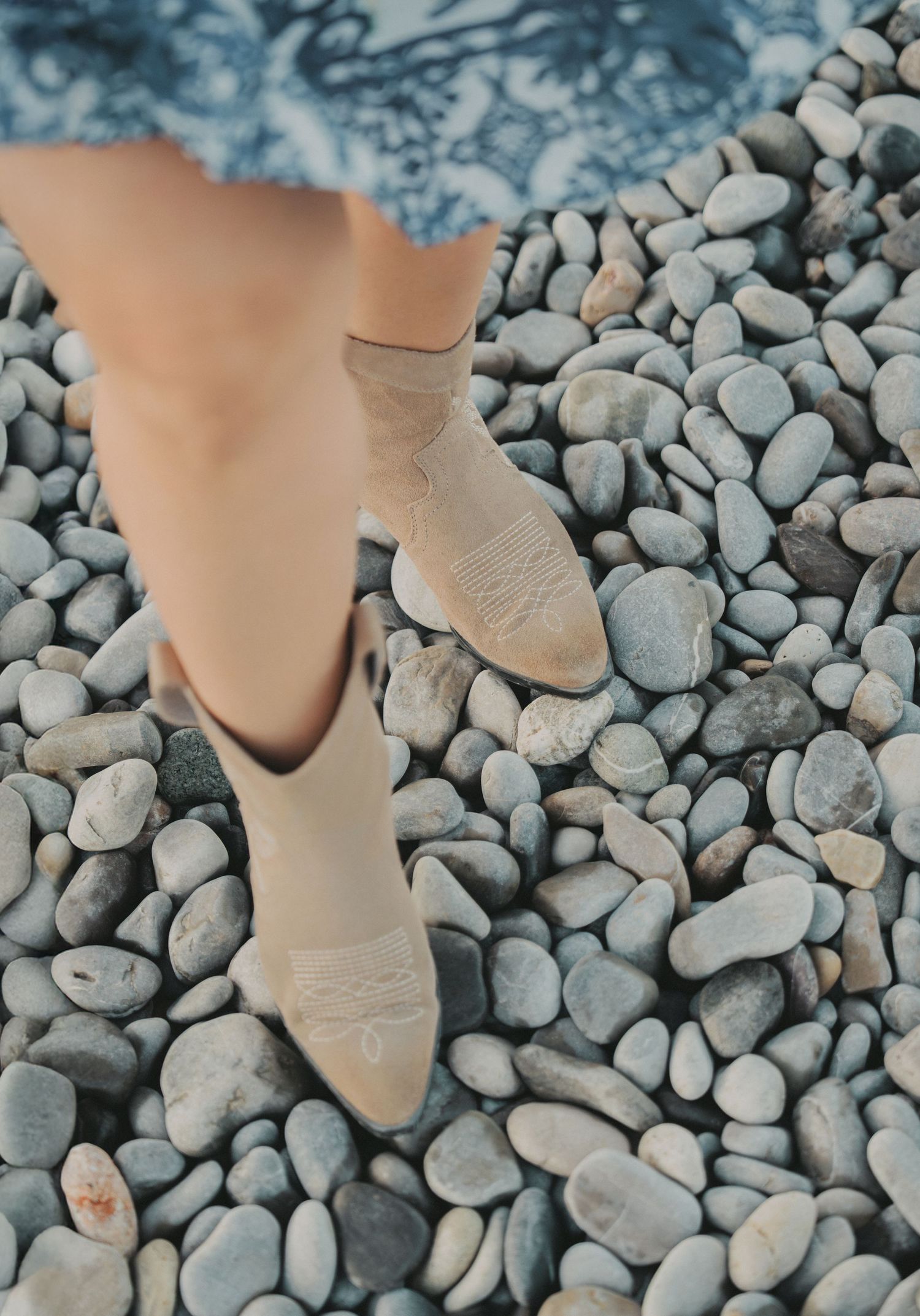 Woman in beige cowboy boots standing on smooth pebble stones at beach.