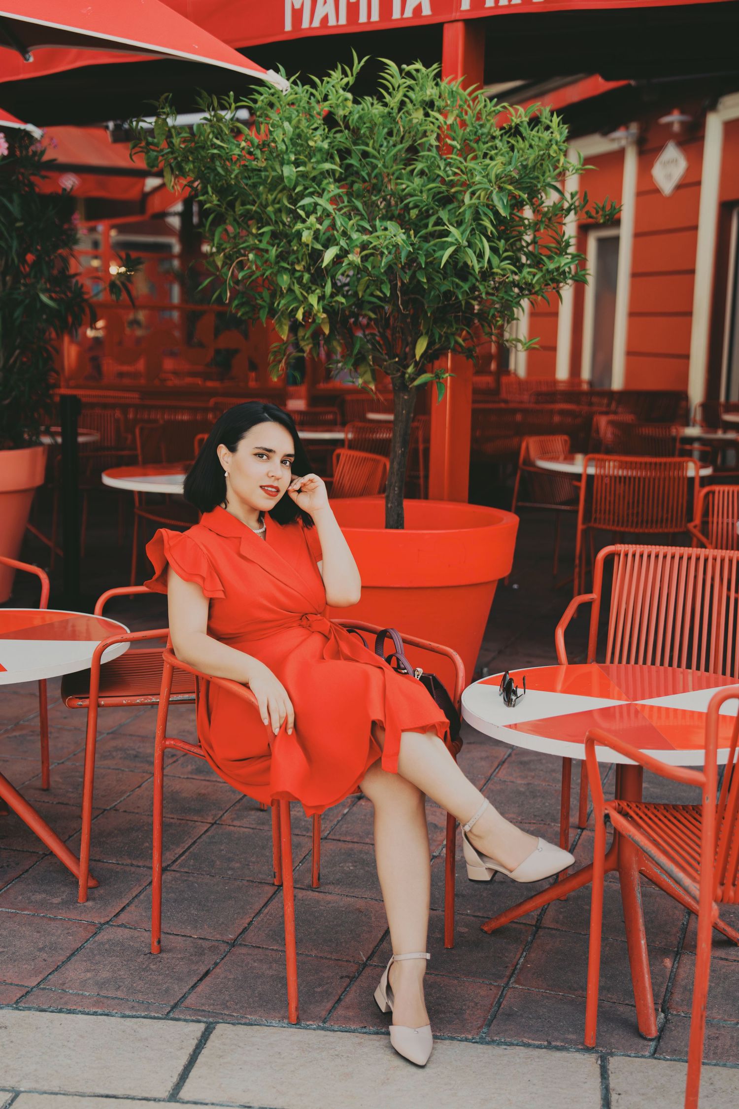 Stylish woman in red midi dress posing at red outdoor restaurant terrace.