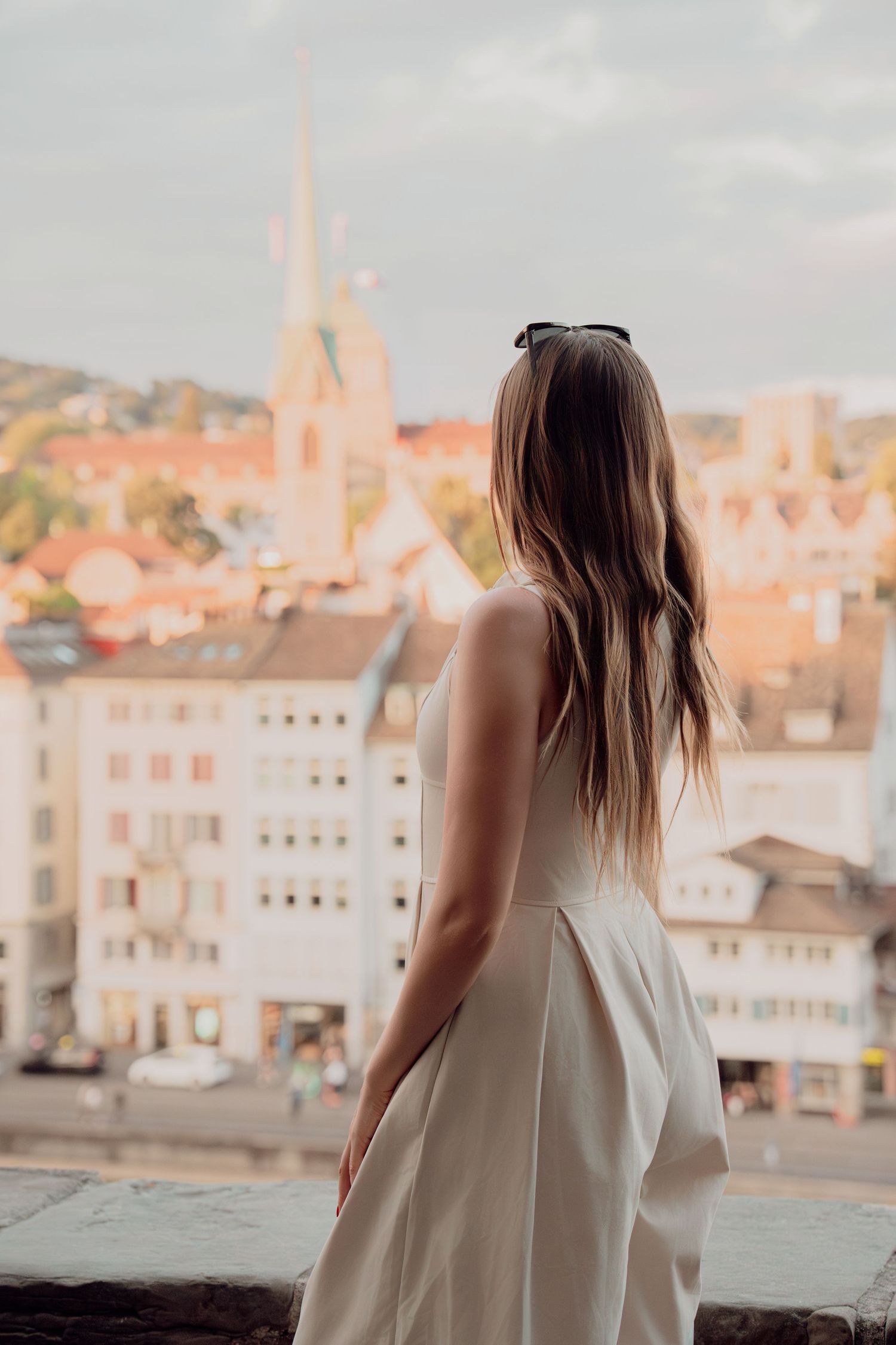 Woman in white dress gazes at Zurich cityscape from elevated viewpoint with church spire visible.