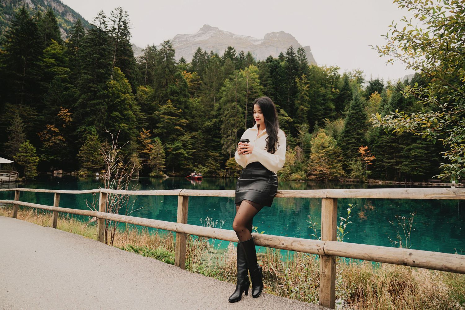 Woman in white sweater and black skirt using phone by wooden fence at stunning turquoise alpine lake.