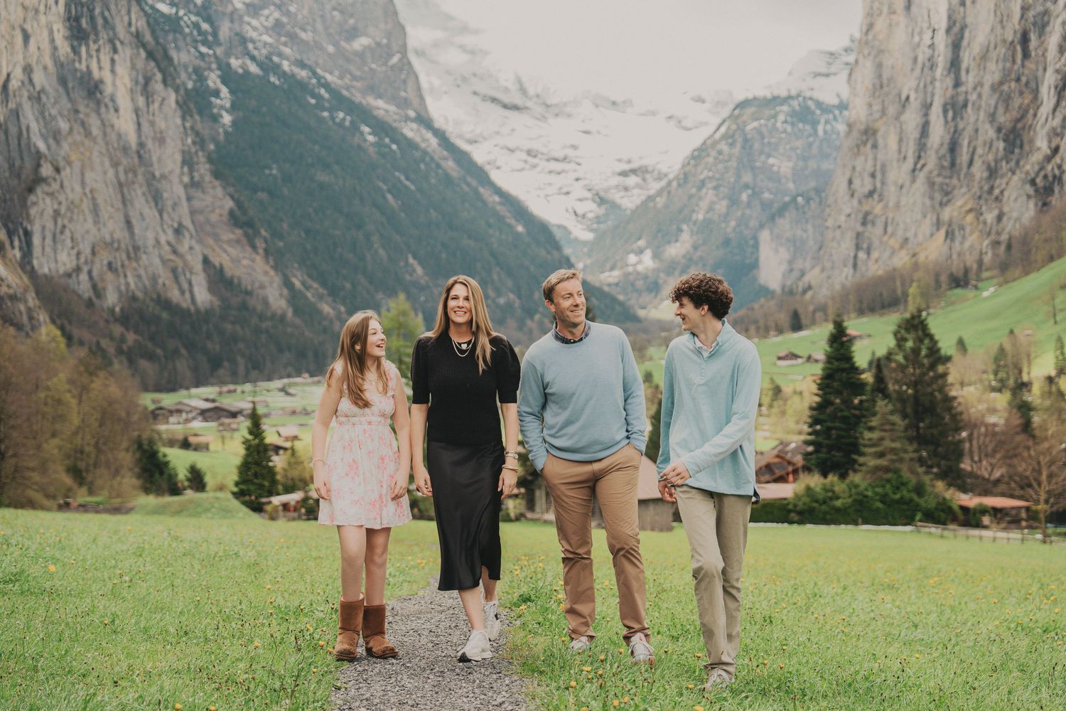 Family of four enjoying a leisurely walk on a grassy path with towering Swiss mountain cliffs behind them.