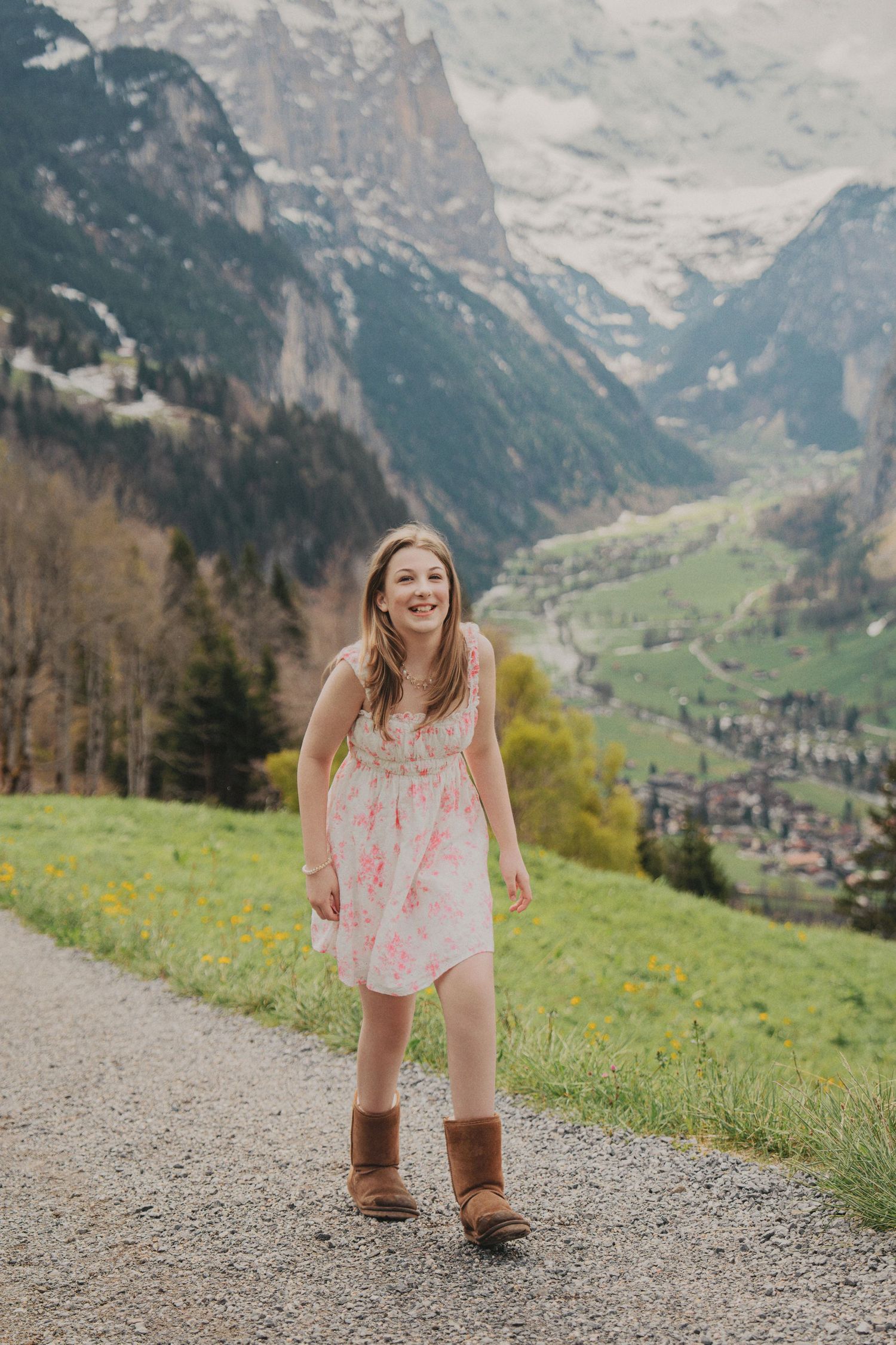 Smiling girl in pink floral dress and brown boots on a gravel path with Alpine valley view.