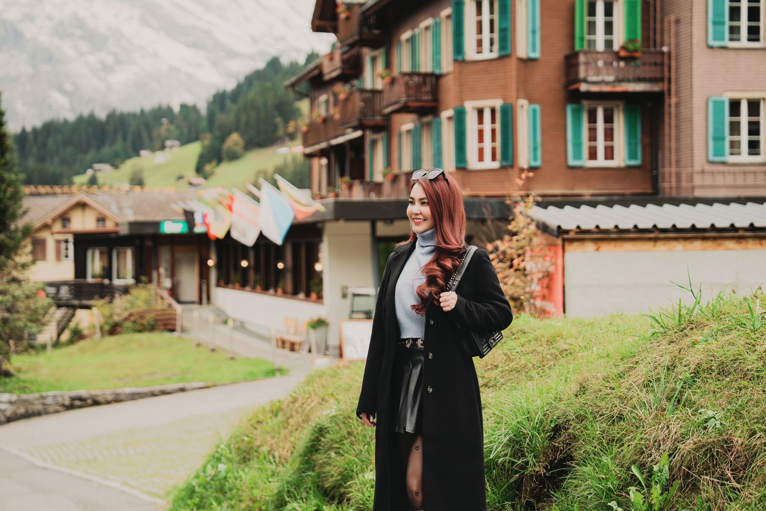 Woman in black coat holds autumn leaves while standing near traditional Swiss Alpine buildings on a cloudy day.