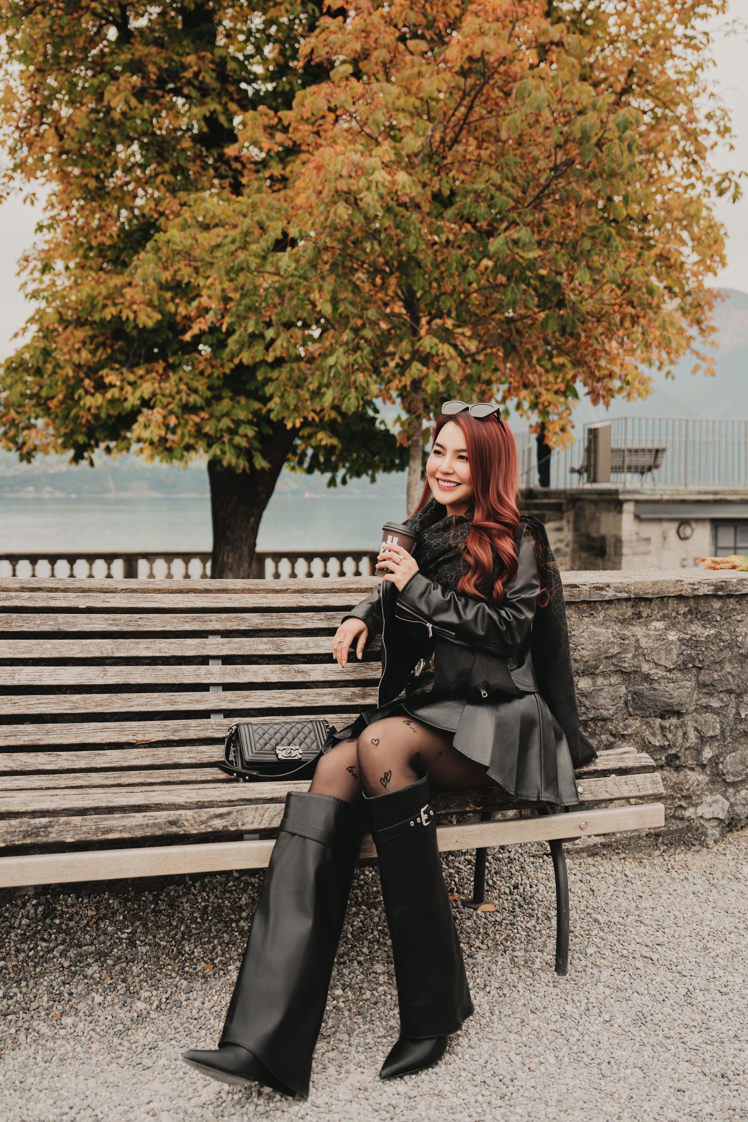Fashionable woman in black leather jacket and knee-high boots posing on bench with autumn tree backdrop.